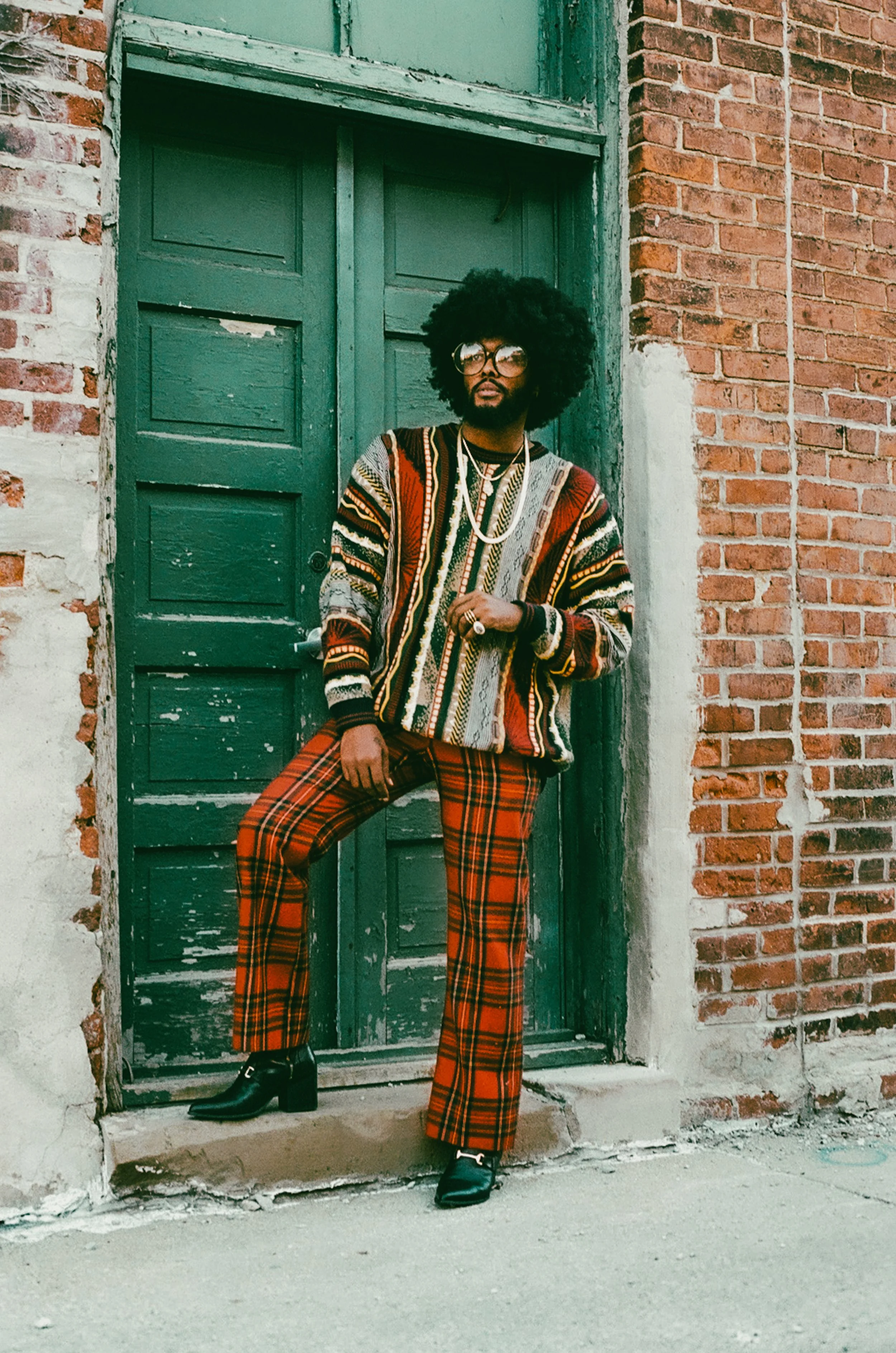A person wearing a colorful sweater and red plaid pants stands against a brick wall and green door, exuding a retro style.