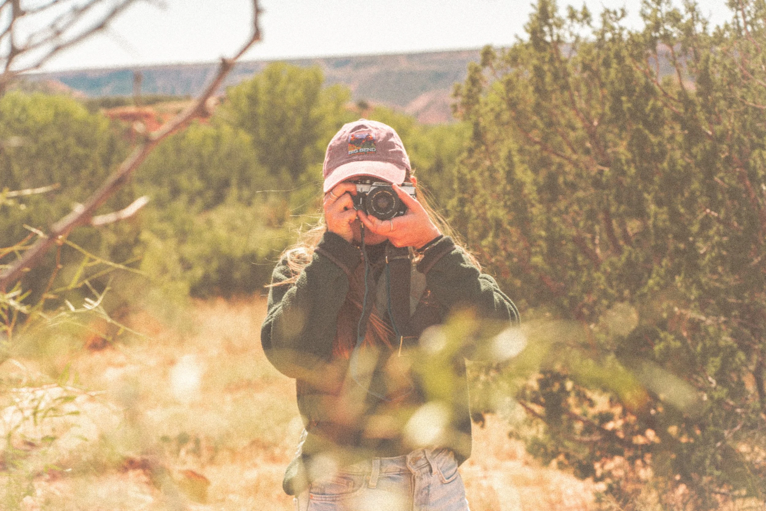 Person in a hat taking a photo with a camera in an outdoor natural setting, surrounded by trees and brush.
