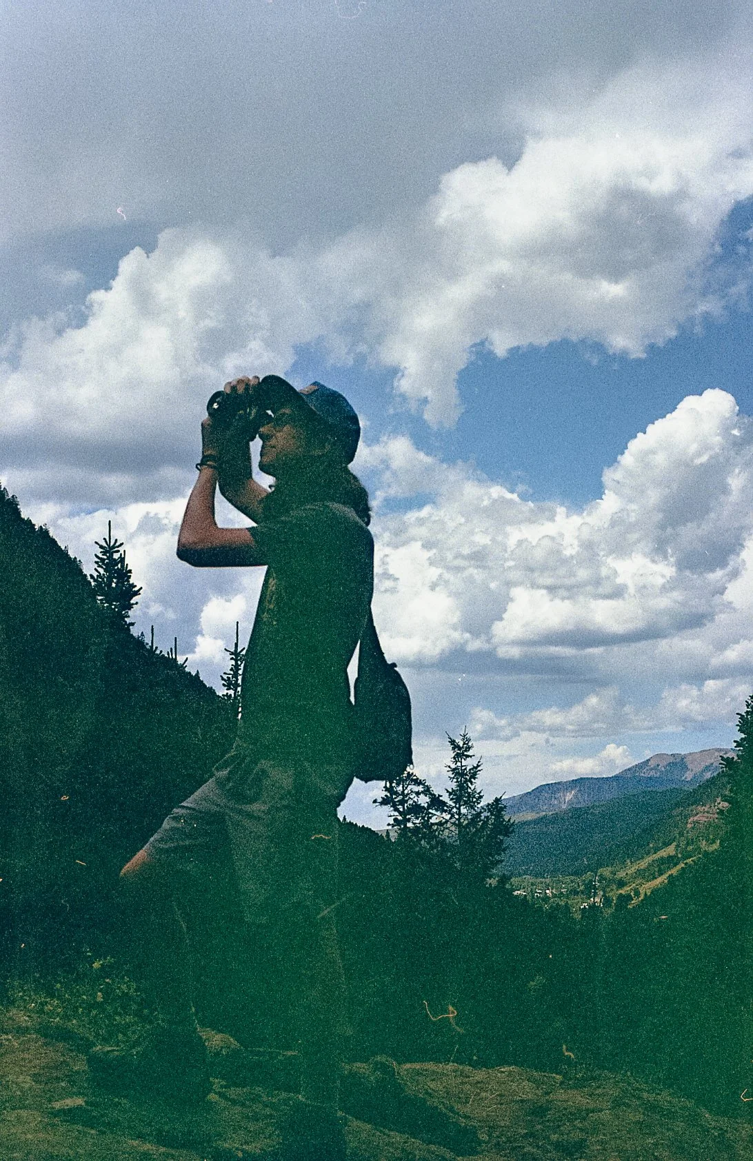 Silhouette of person hiking with binoculars and backpack in a mountainous landscape, cloudy sky above.