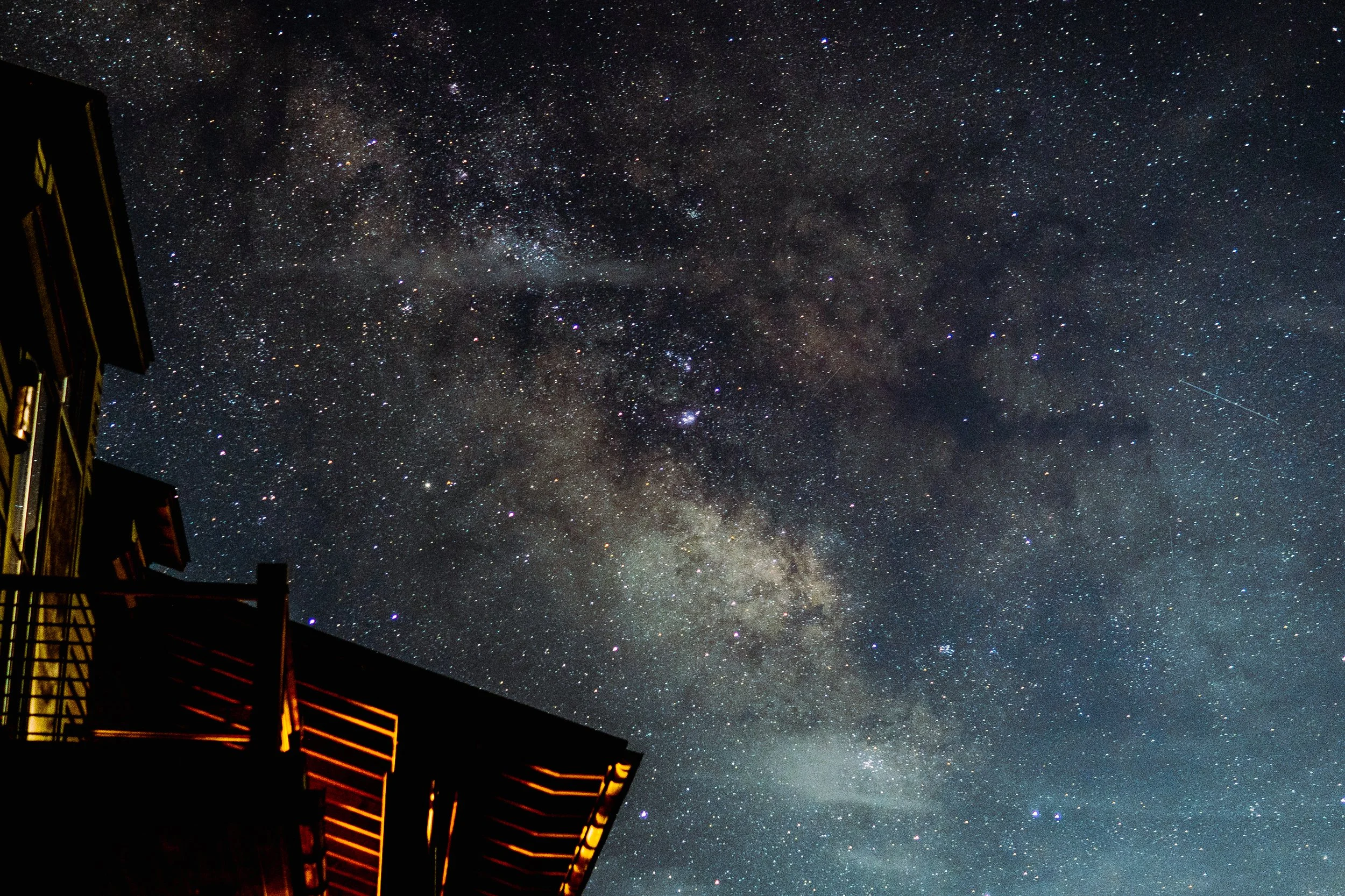 Night sky with starry Milky Way galaxy and part of a house with illuminated windows.