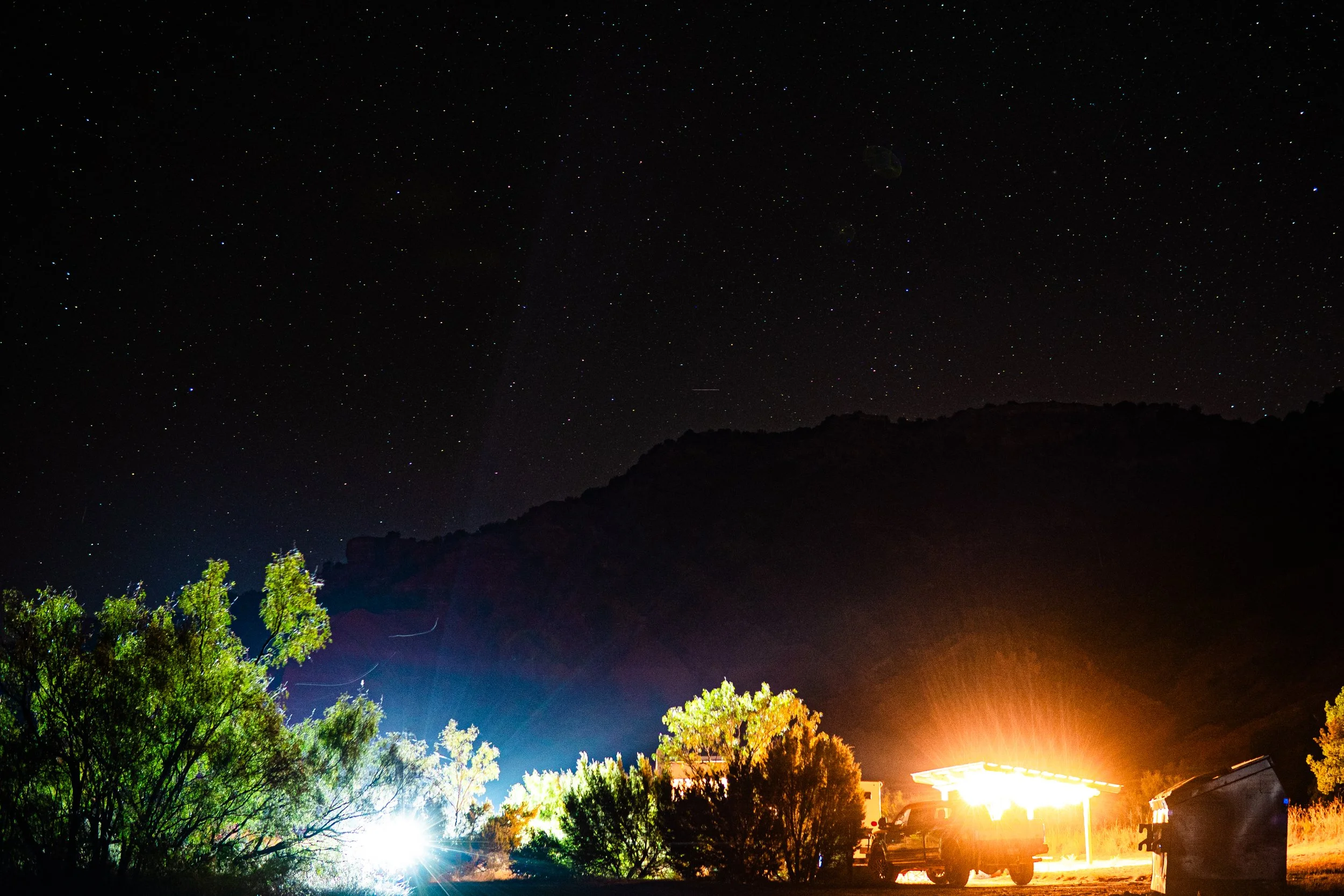 Night landscape with starry sky, illuminated trees, a pickup truck under a bright light, and a silhouetted mountain.