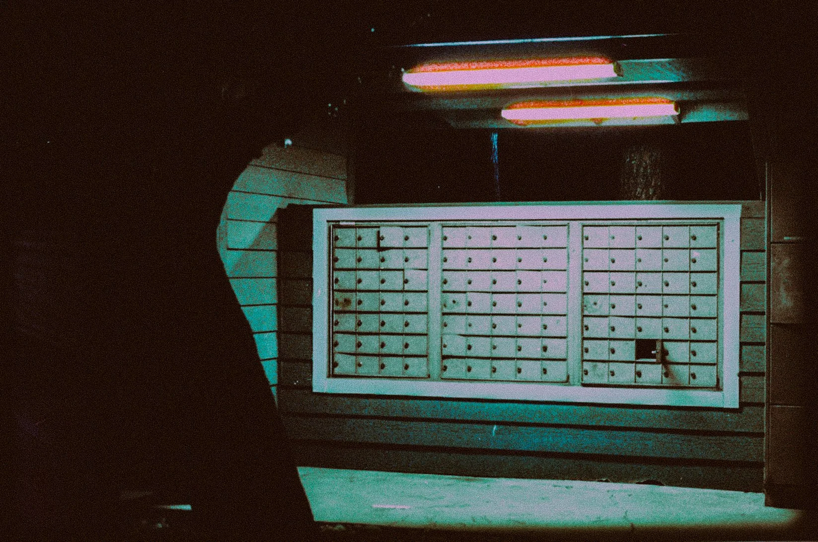 A set of outdoor mailboxes under dim lighting, with one door open, against a wooden wall.