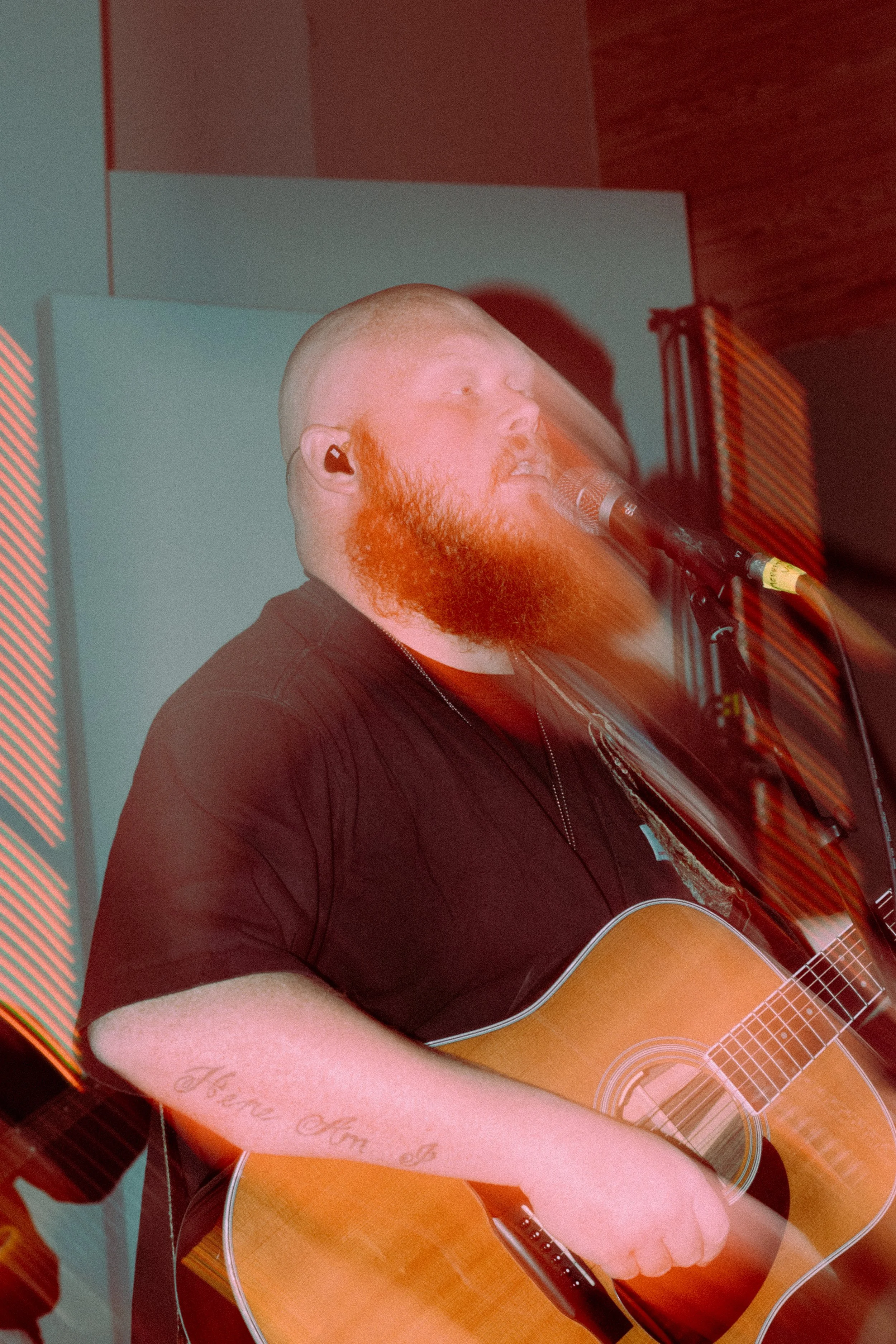 Musician with a beard playing acoustic guitar and singing into a microphone, wearing a black shirt on stage, with blurred motion effect.