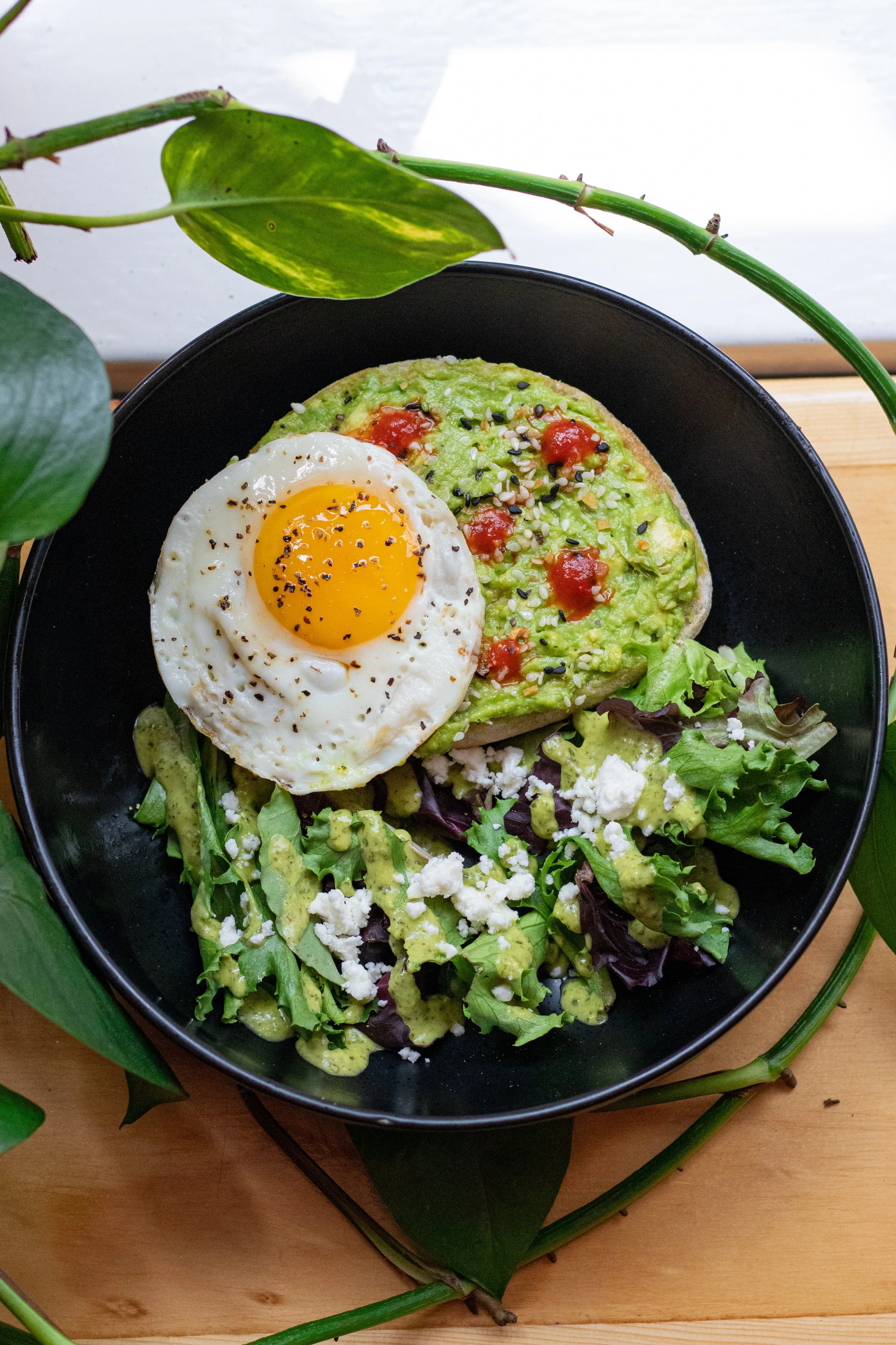 Black plate with avocado toast topped with a fried egg and sriracha; side salad with feta cheese and green dressing; surrounded by green leaves.