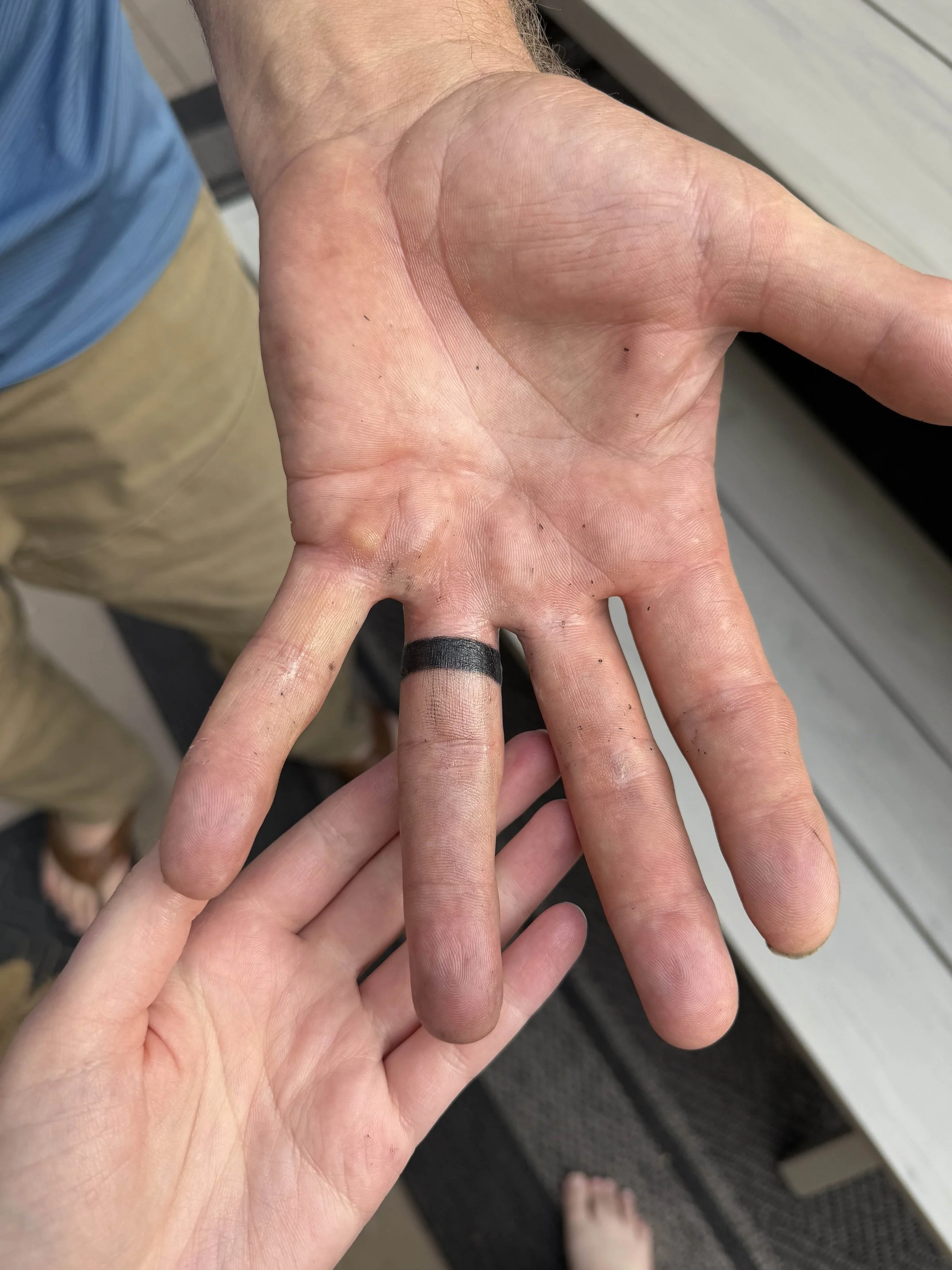 A person with a tan hand has a black ring on the pinky finger, with a hand resting on another person's hand. The background shows part of a gray and wood surface.
