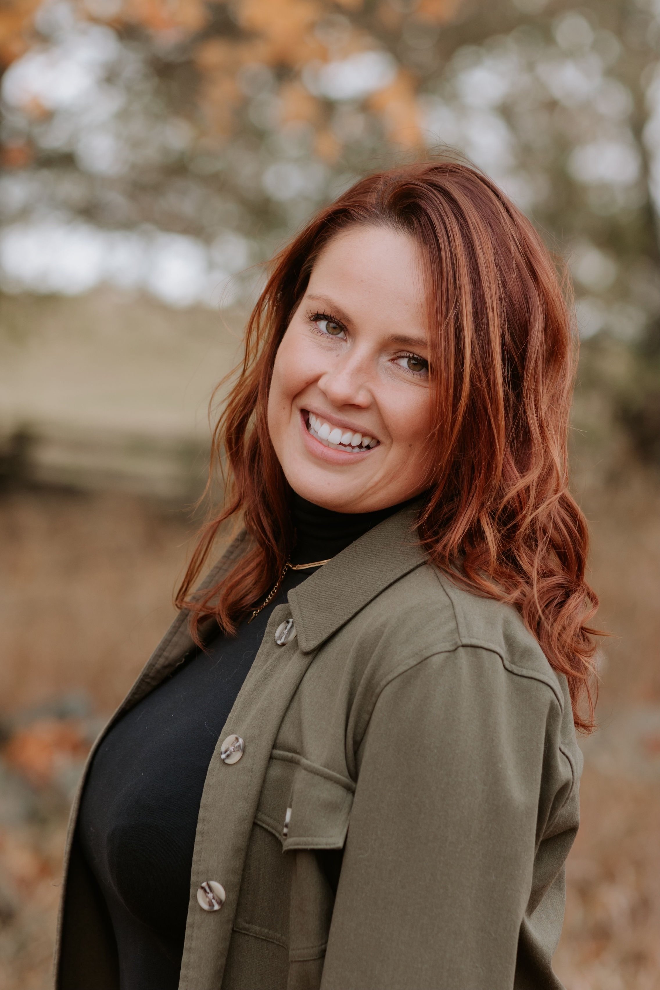 A woman with reddish hair smiling outdoors, wearing a black top and an olive green jacket.