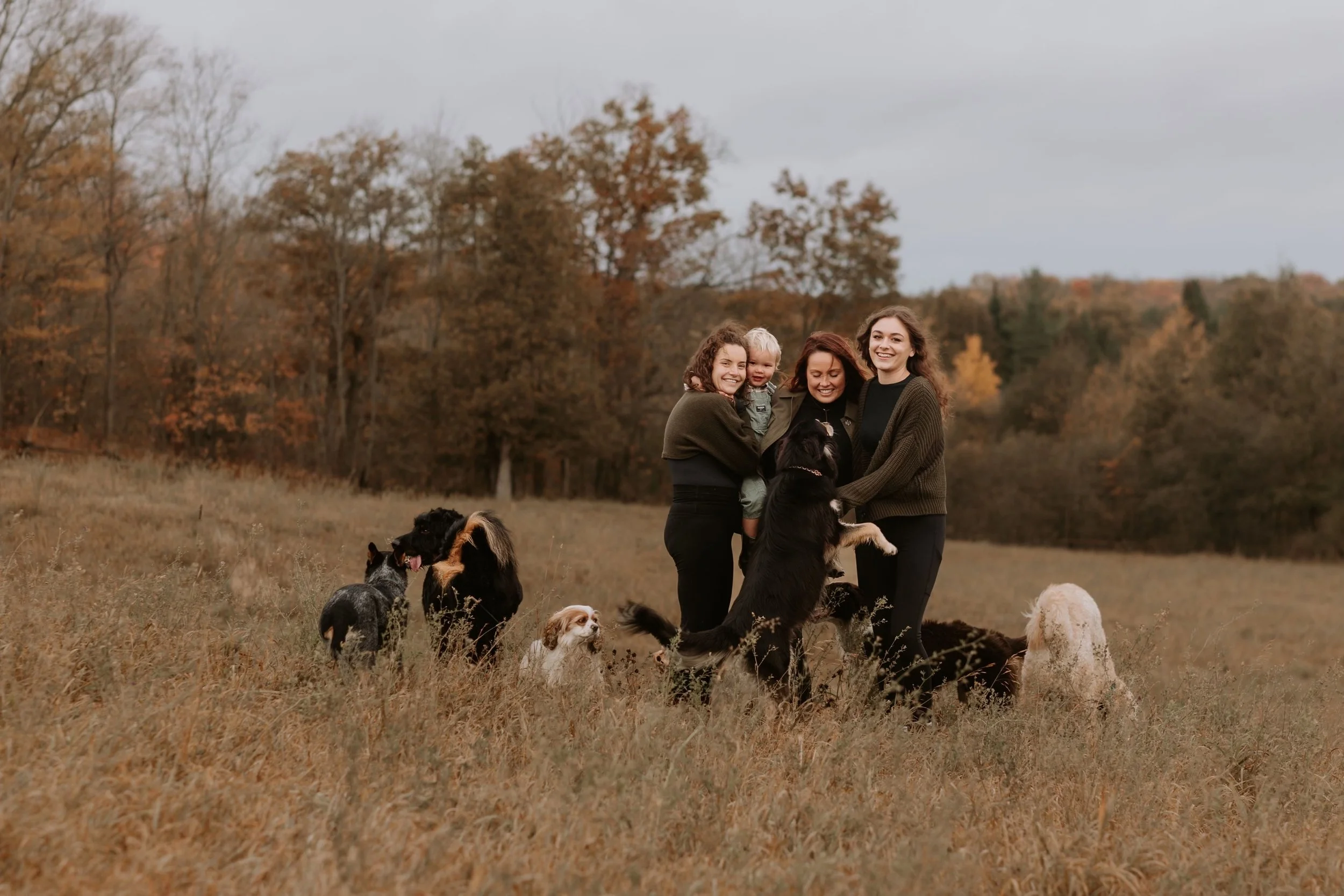 Three women and a young girl standing in a grassy field with several dogs, with autumn trees in the background.