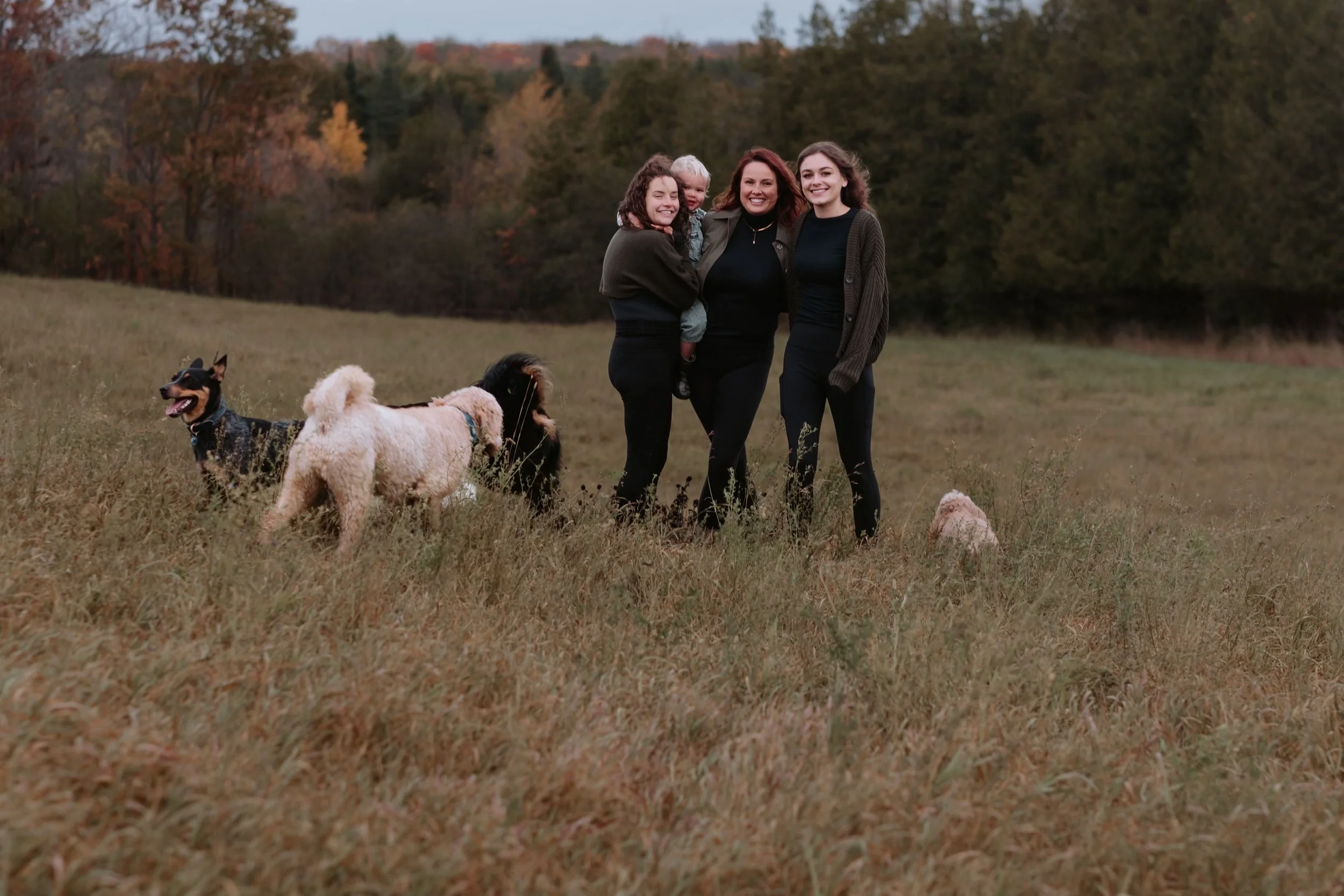 Four women and four dogs standing in a grassy field with trees in the background, during autumn.