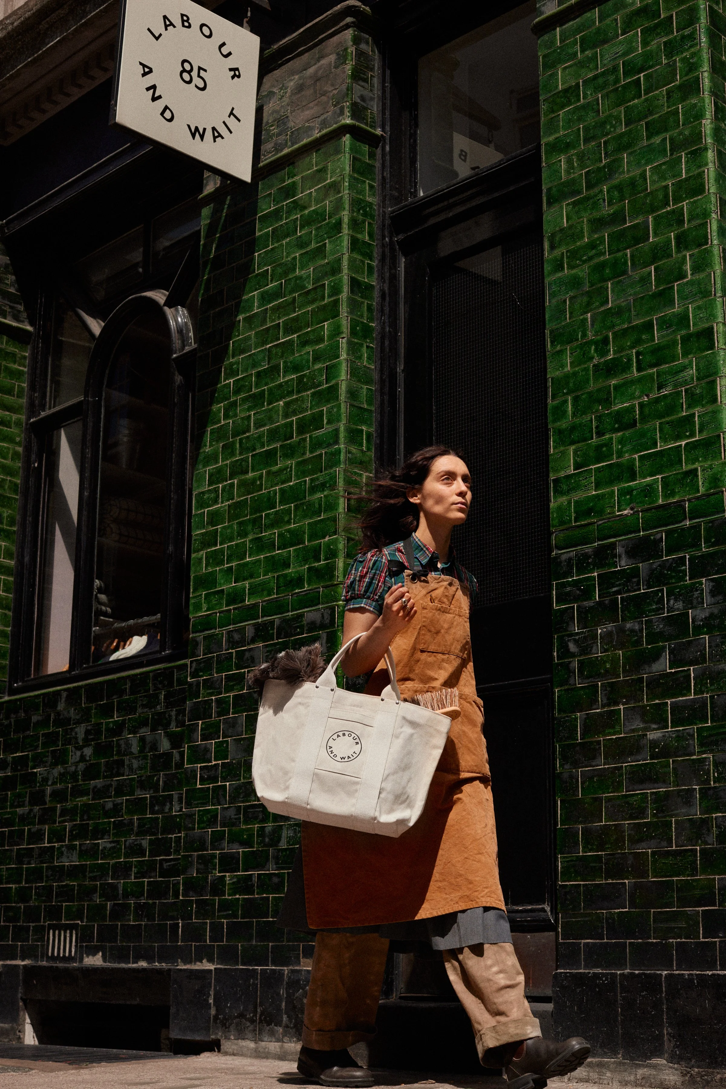 A woman with dark hair walking past a dark green tiled building, carrying a large beige tote bag that has 'Labour and Wait' printed on it, wearing a plaid shirt with rolled-up sleeves, a brown apron, loose pants, and dark shoes.