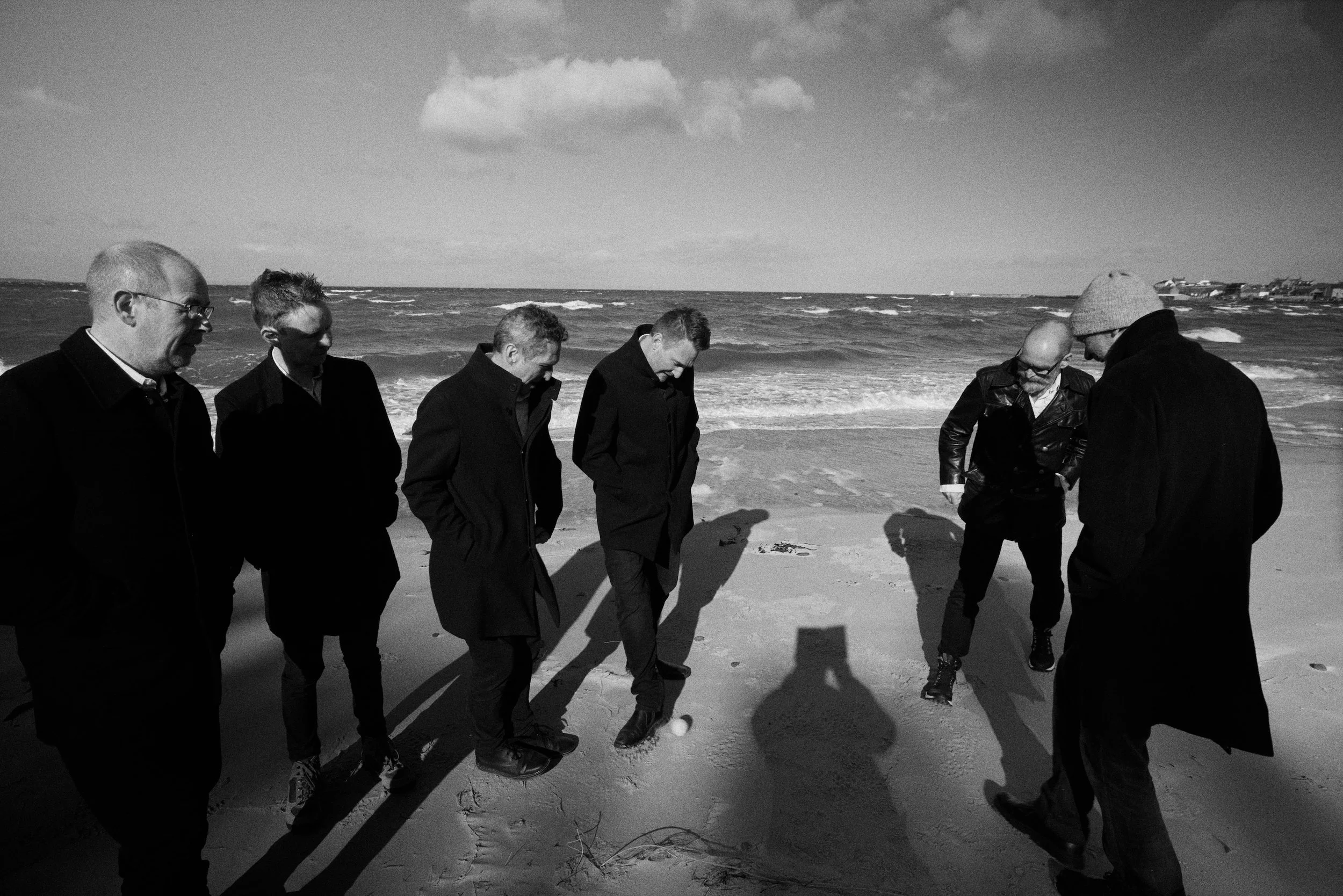 Seven men standing on a beach in winter coats, looking at something in the sand with the ocean in the background on a cloudy day.