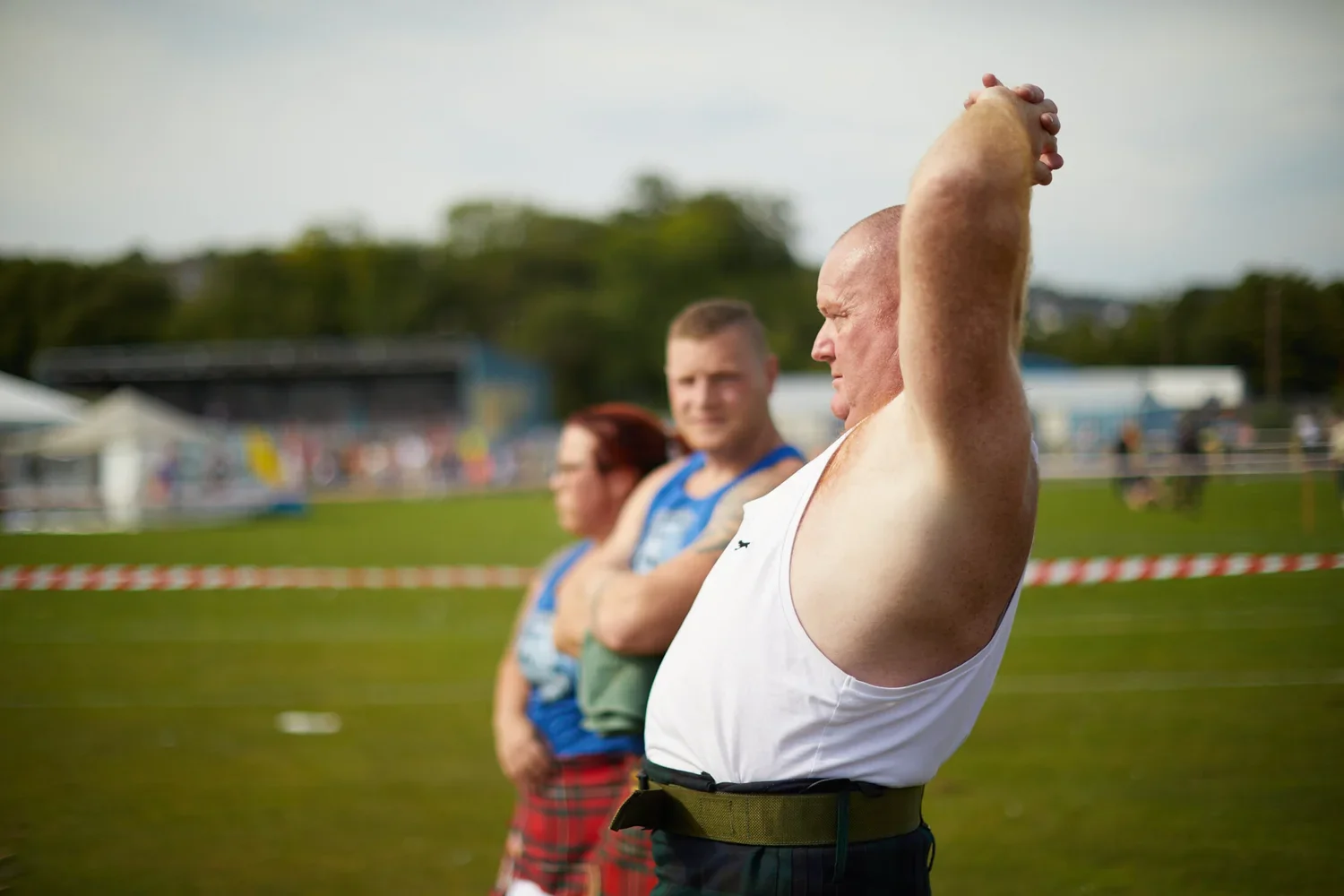 Three athletes preparing for a shot put event at an outdoor track and field competition, with a grassy field and spectators in the background.