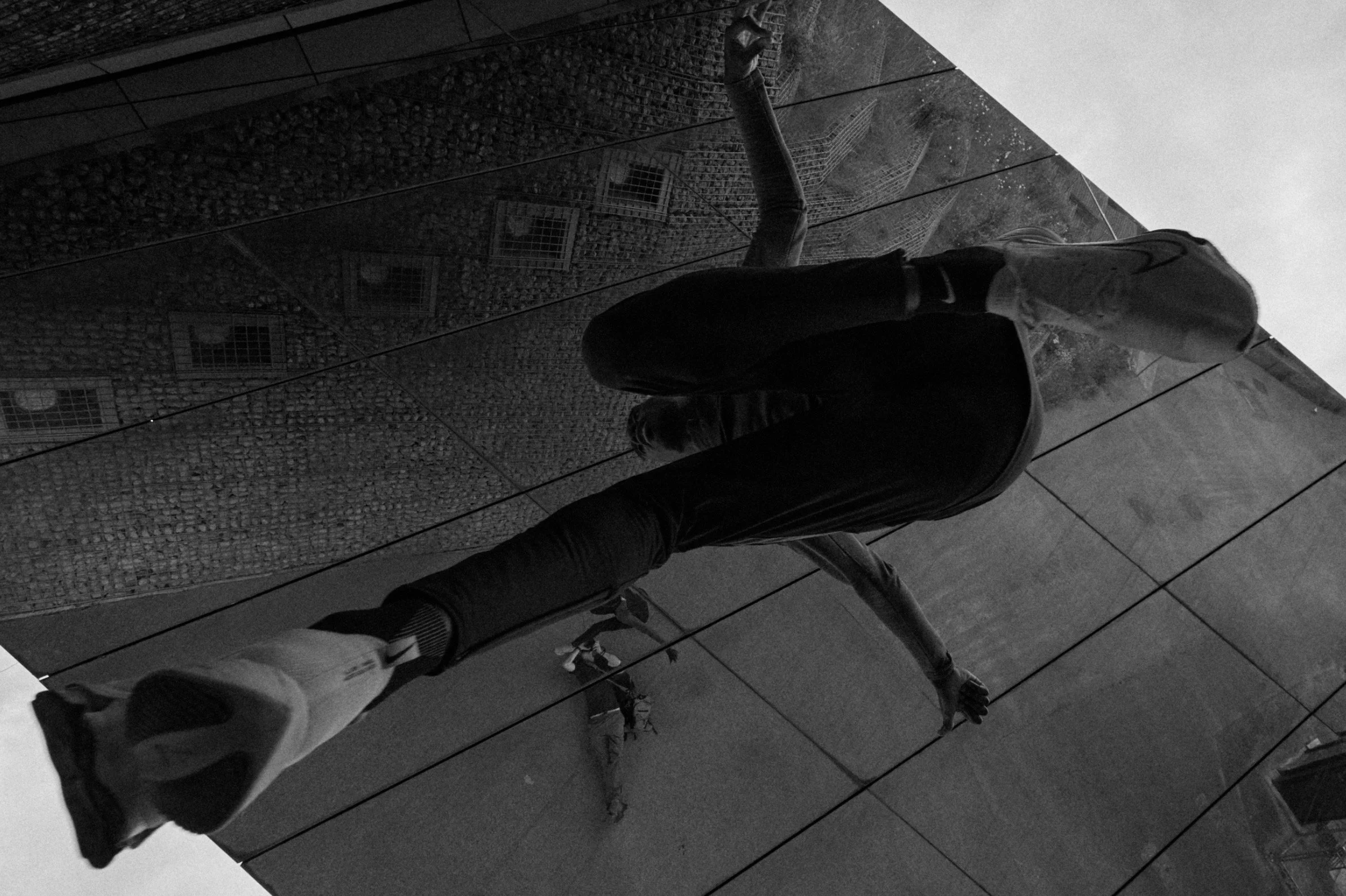 A person in black clothing balancing on a narrow ledge or mirror with their right hand touching the surface, reflected in a large mirror or glass wall showing a textured brick wall and a person taking a photo in the background.