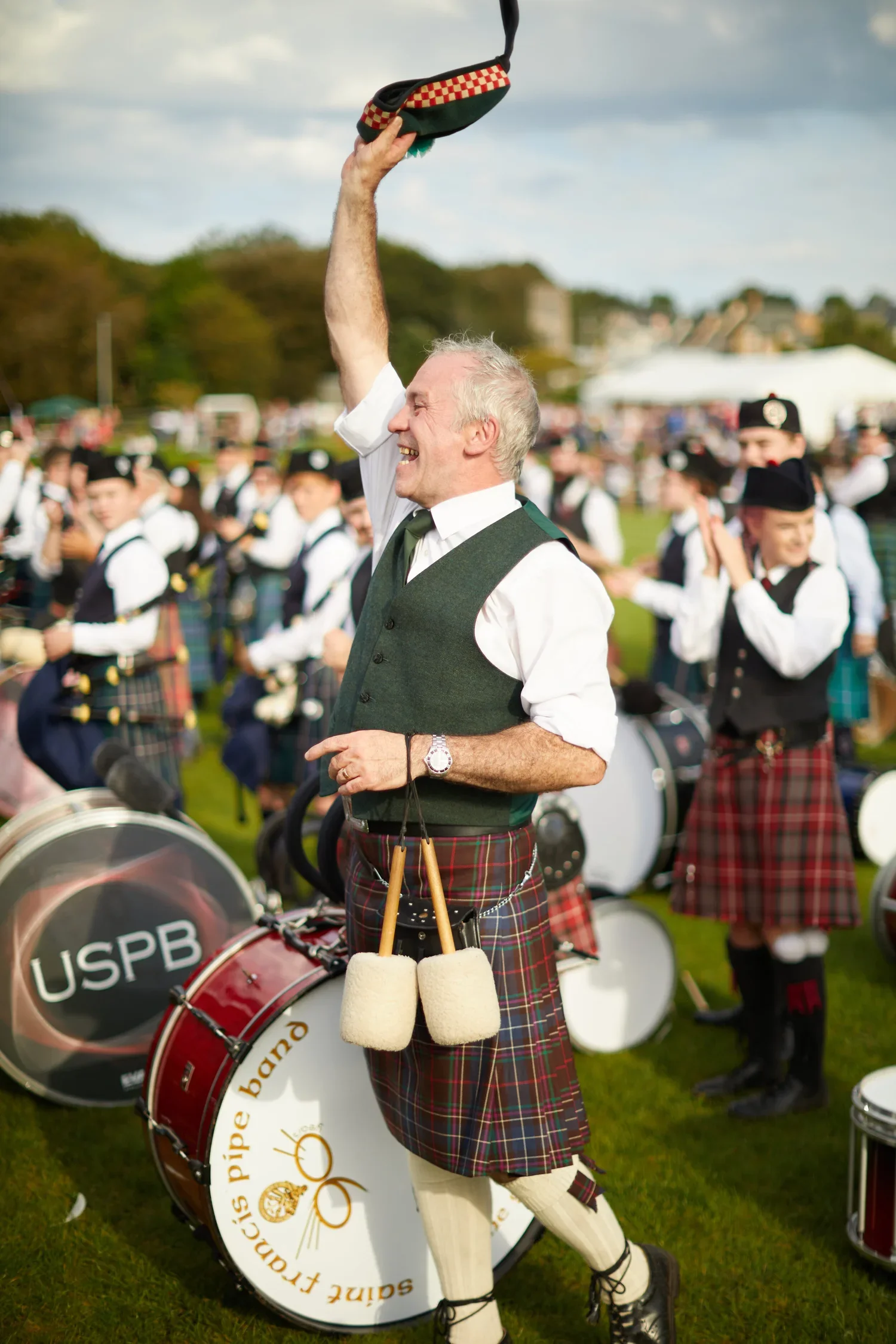 A man in traditional Scottish attire, including a kilt, raising a hat and smiling at a gathering of bagpipe players in a field.