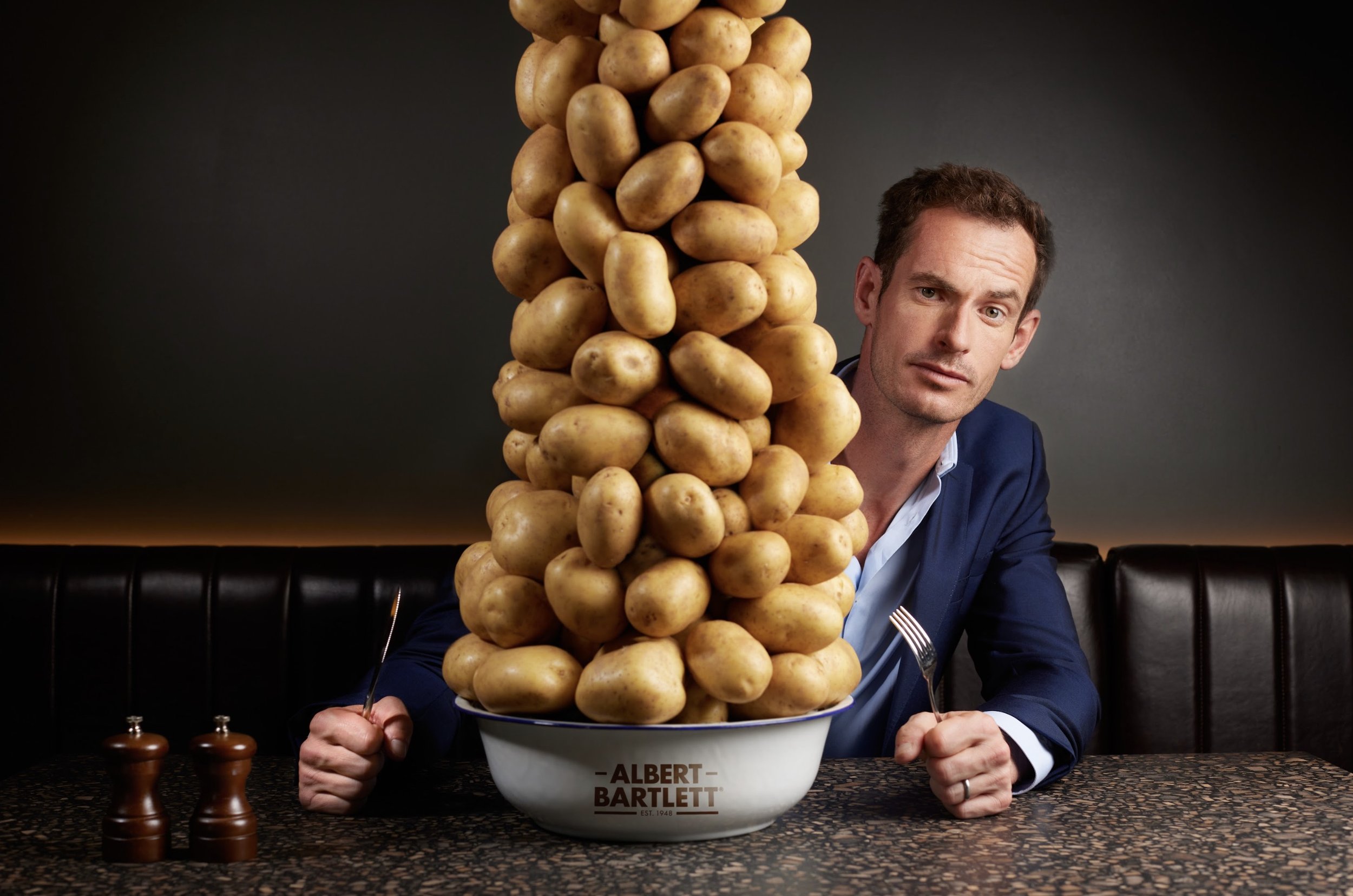 A man in a suit sitting at a table with a large bowl of potatoes and a tower of potatoes rising above the bowl, with salt and pepper shakers on the table.