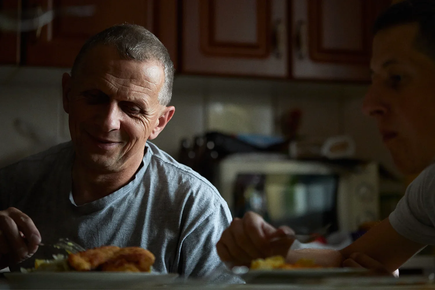 A smiling man with short gray hair sitting at a kitchen table, serving food, with another person partly visible on the right side.