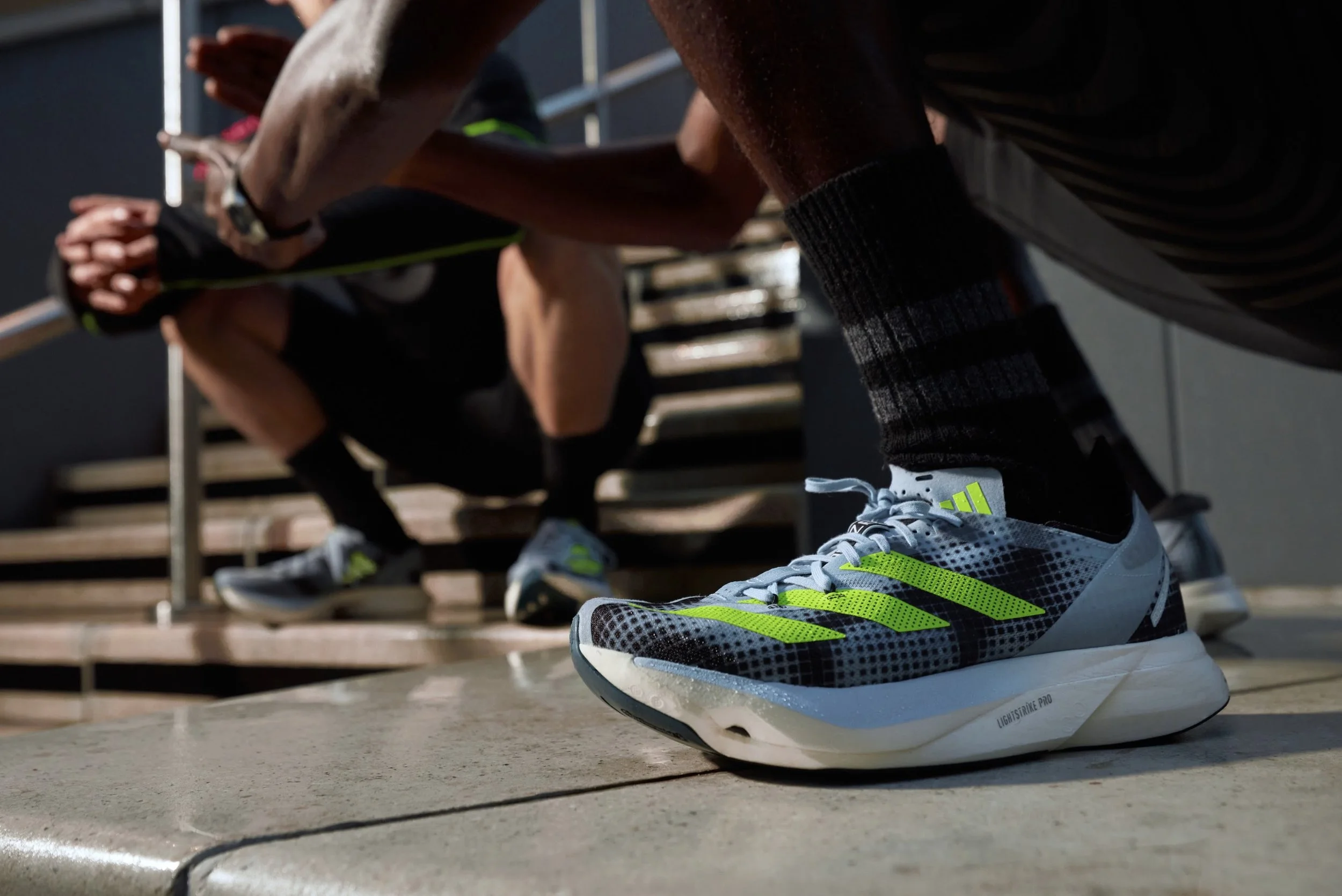 Close-up of a person's running shoe with bright yellow stripes on the sole, worn by someone preparing to sprint during a workout or race, with other runners in the background.