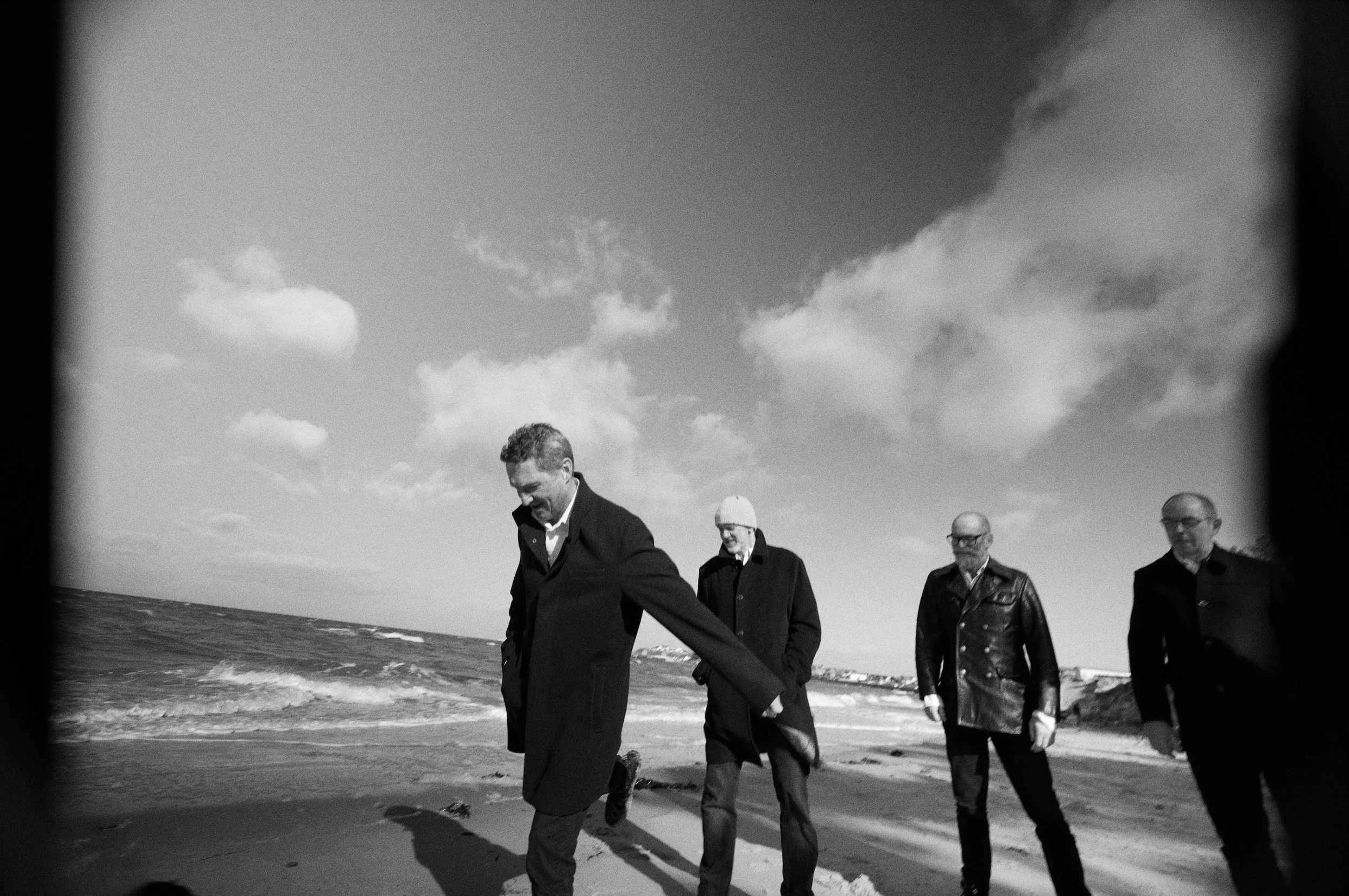 Four men walking along a beach in coat and sunglasses, with waves in the background and a cloudy sky.