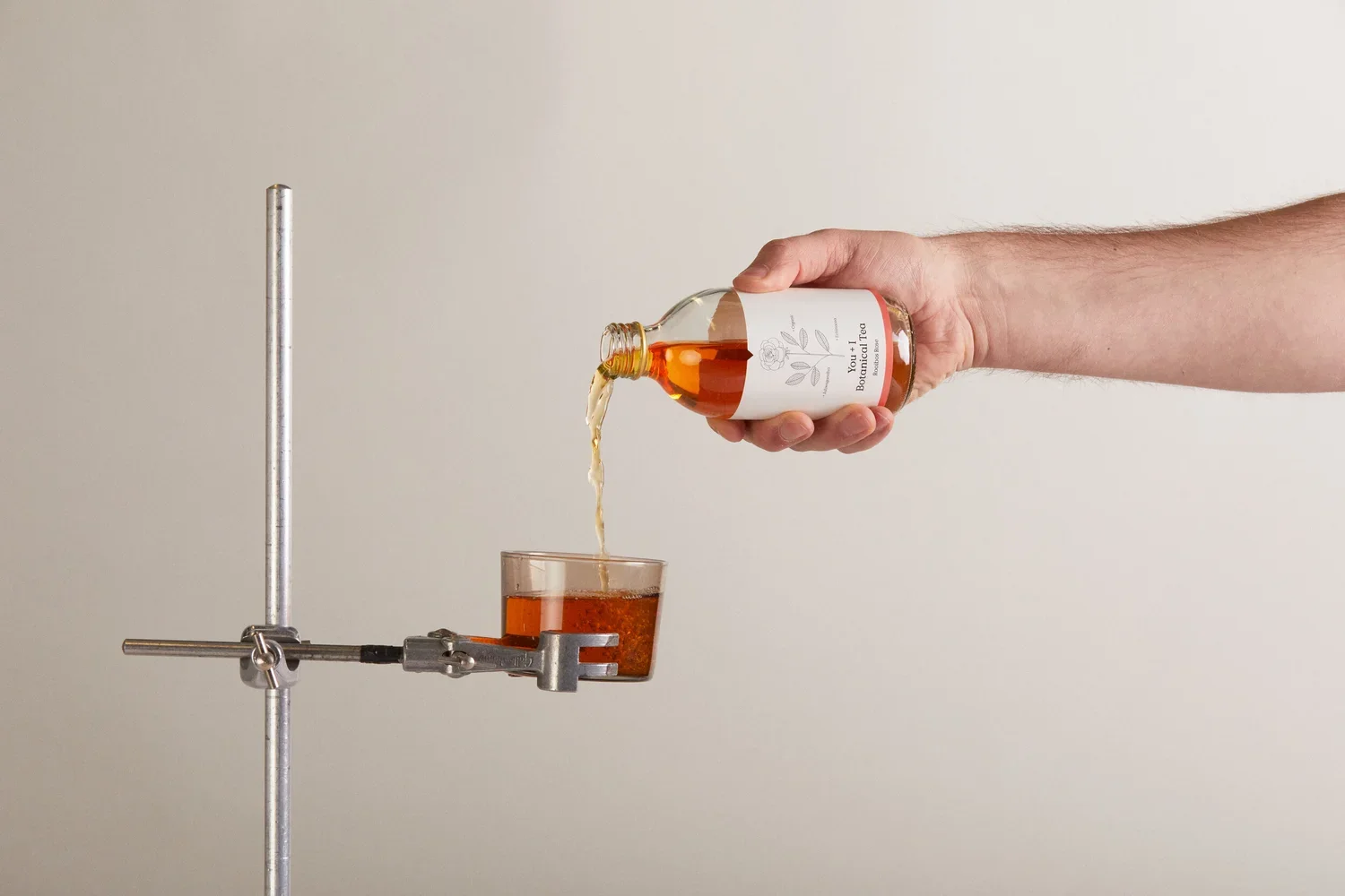 Person pouring tea from a bottle into a glass placed on a lab stand.