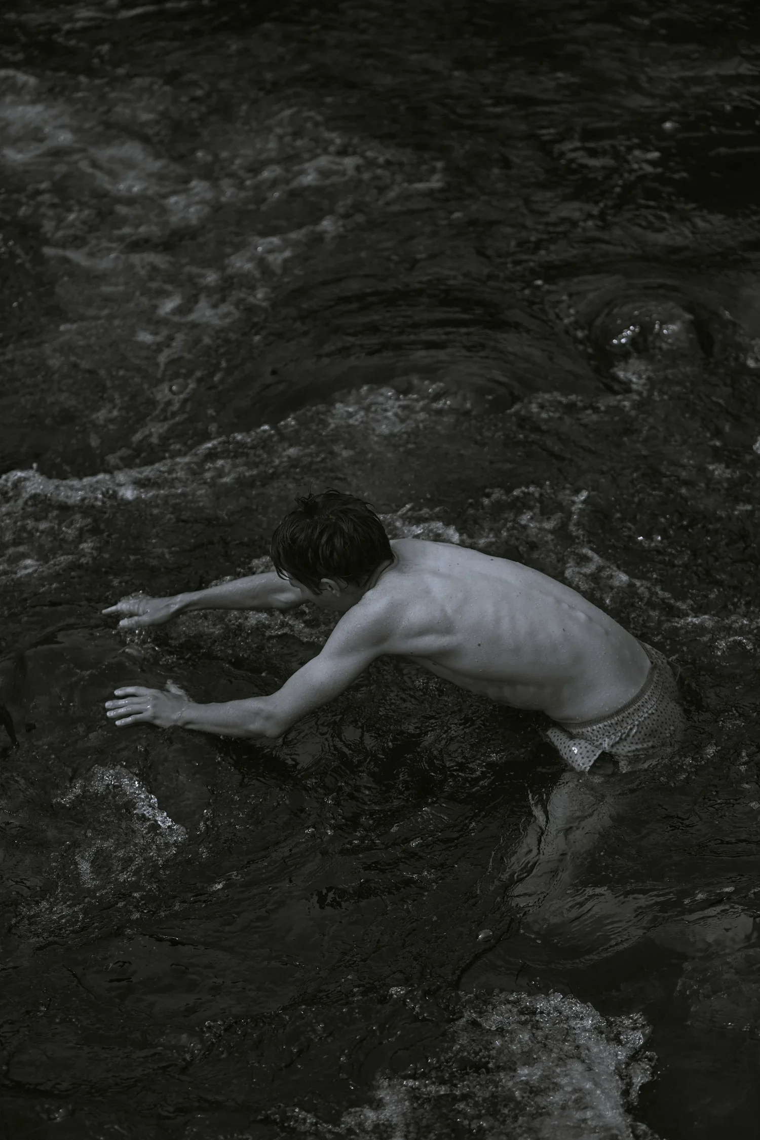 A shirtless boy in patterned swim trunks reaching for a rock in a dark river.