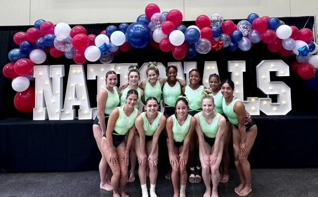 Group of nine women in light green leotards posing in front of a backdrop with balloons and white marquee letters spelling NAYLIS.