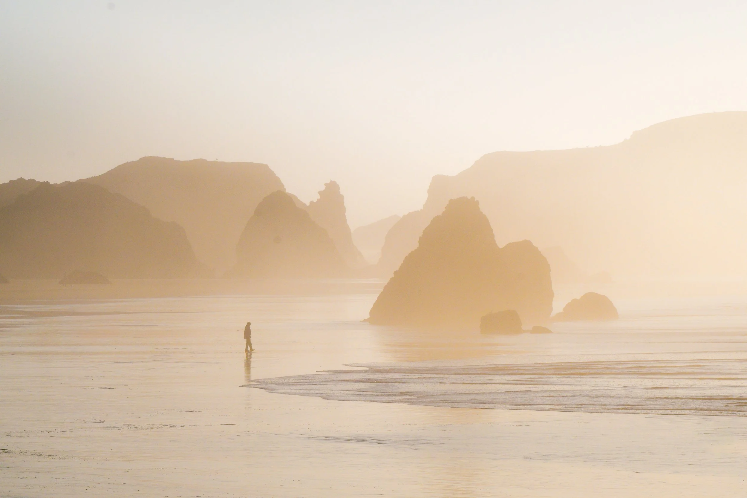 A person walking along a misty Oregon beach with large rocks and cliffs in the background at sunrise or sunset.