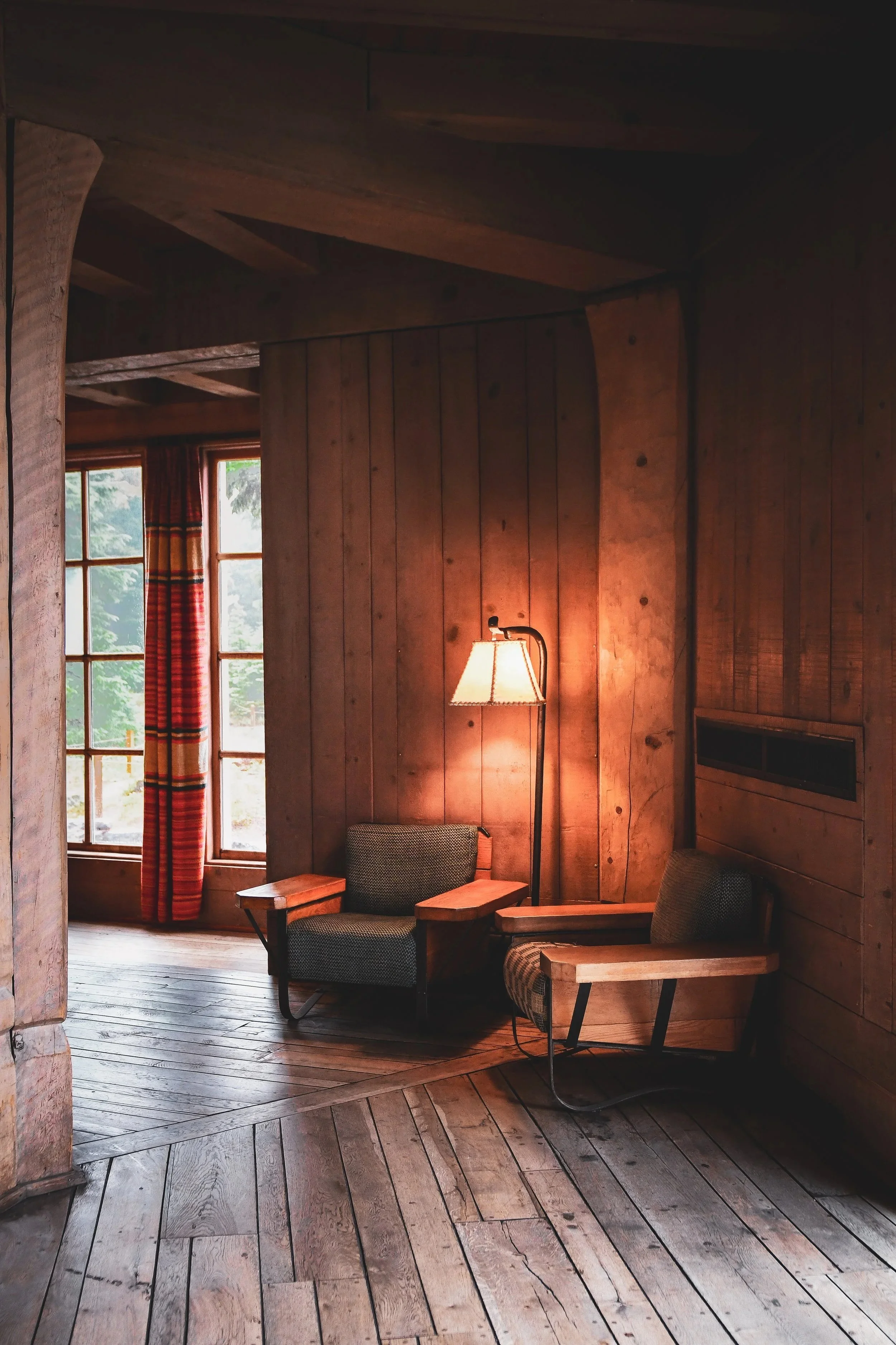 A cozy corner of a room in Oregon's Timberline Lodge with wooden panel walls, featuring two mid-century modern armchairs with wooden arms, a standing lamp between them, a window with red plaid curtains, and a rustic wooden floor.