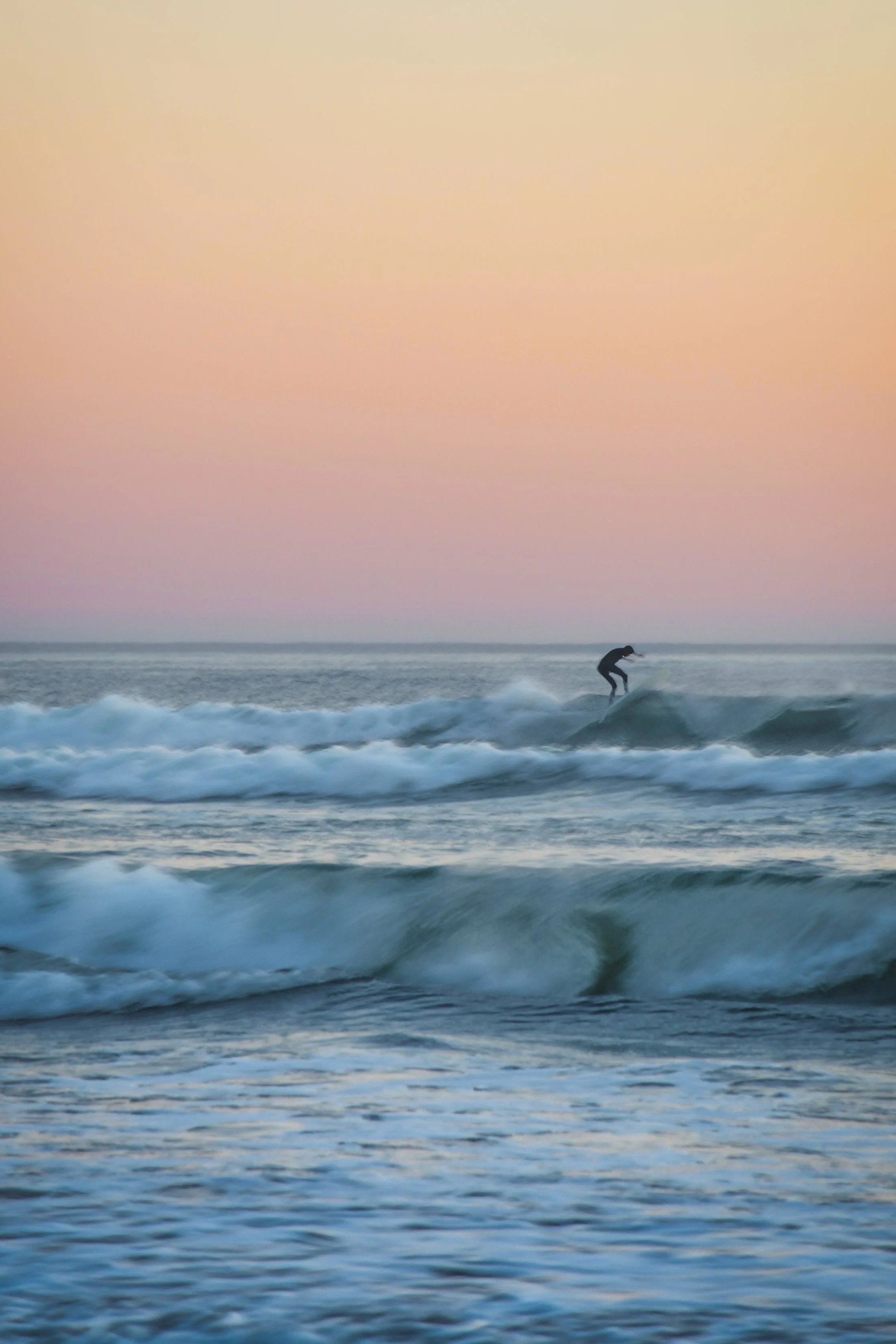 A person surfing on a wave in the Pacific ocean during sunset with a pastel-colored sky, near Cannon Beach,