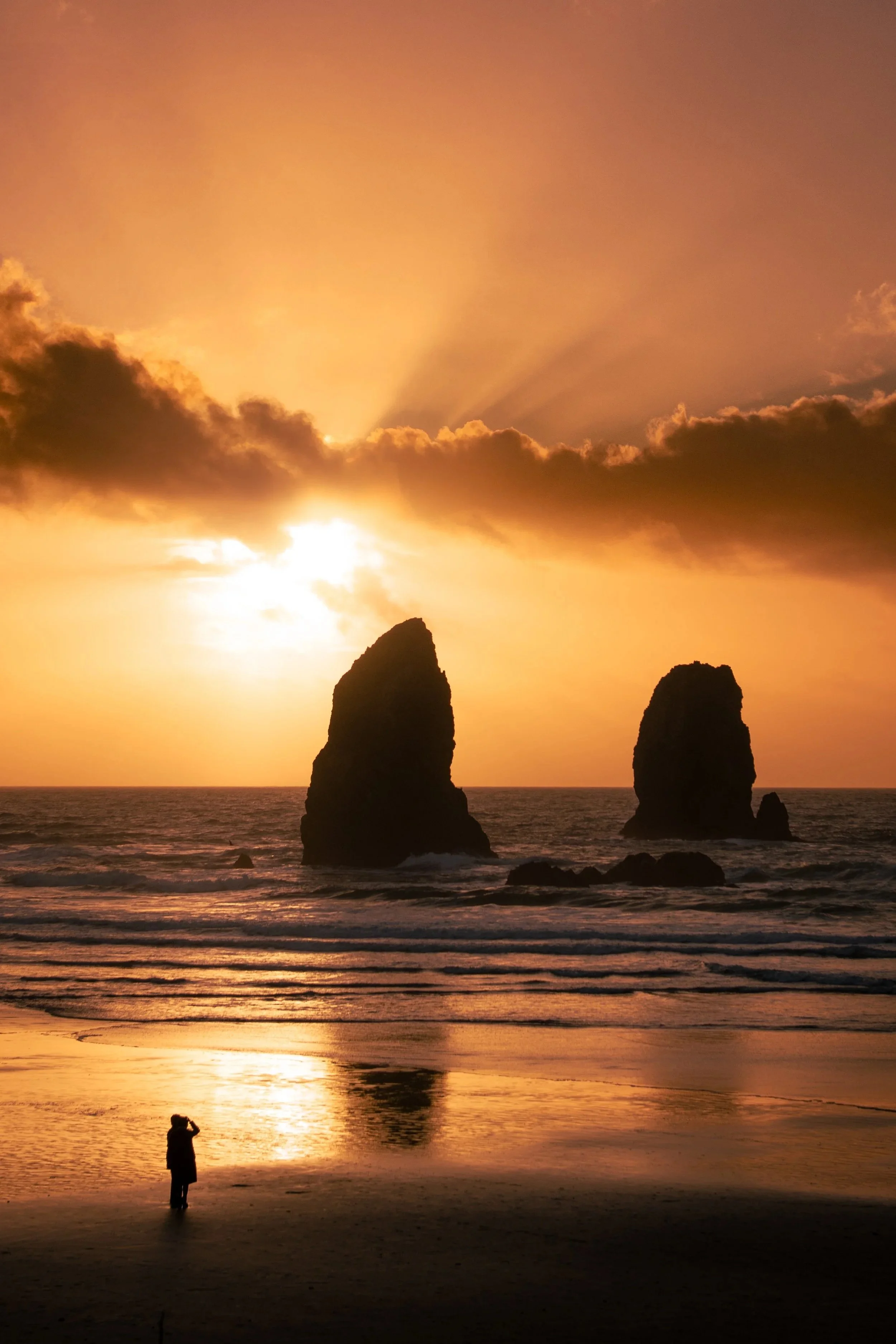 Silhouetted person standing at Cannon beach at sunset with two large sea stacks in the ocean and a colorful sky with clouds