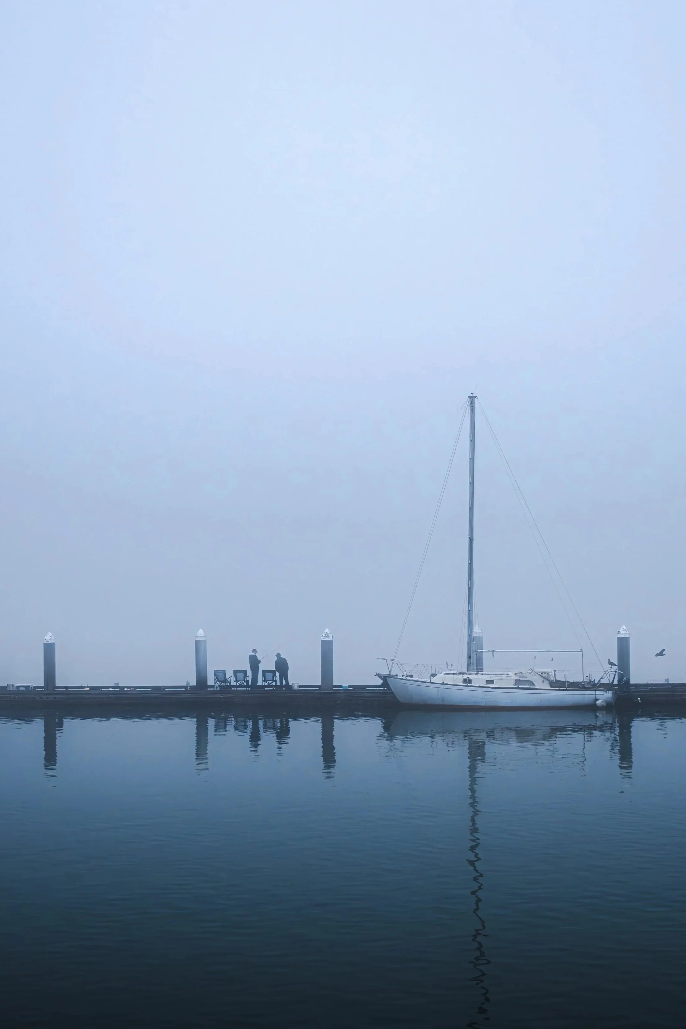 A foggy scene at the Bandon, Oregon marina with a sailboat docked at the pier, two people standing and talking, and a seagull flying nearby.
