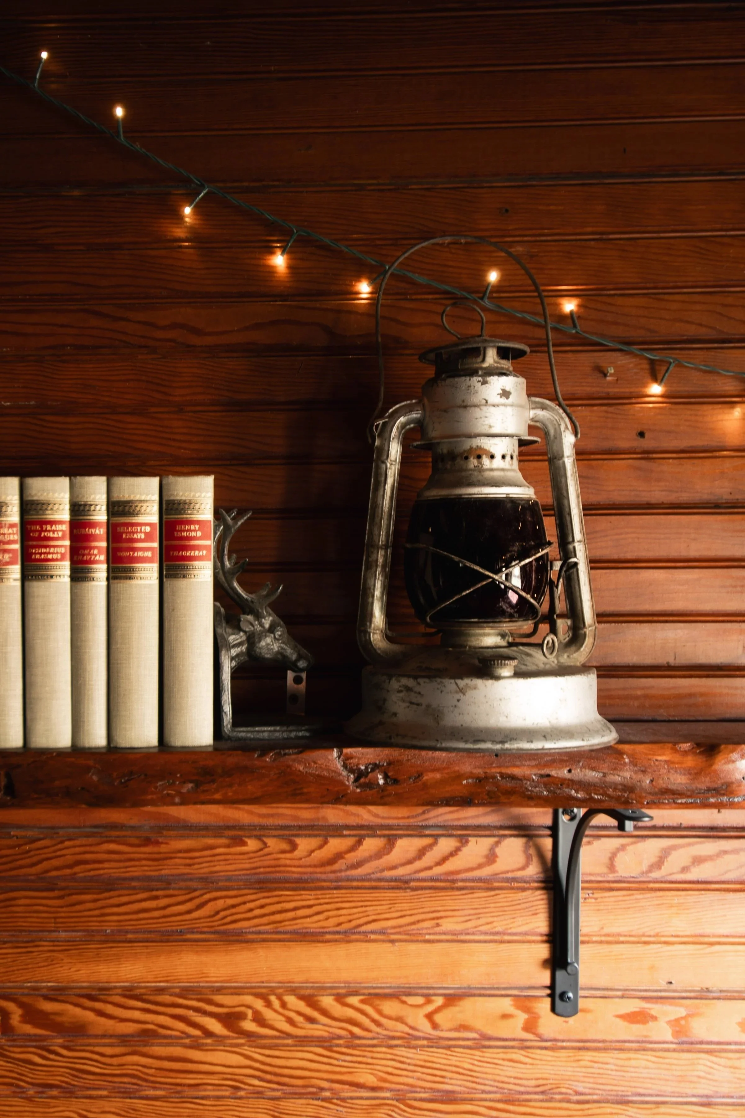 A vintage lantern on a wooden shelf next to a grey deer figurine and a row of seven books with cream covers and red labels, set against a wooden paneled wall with string lights overhead.