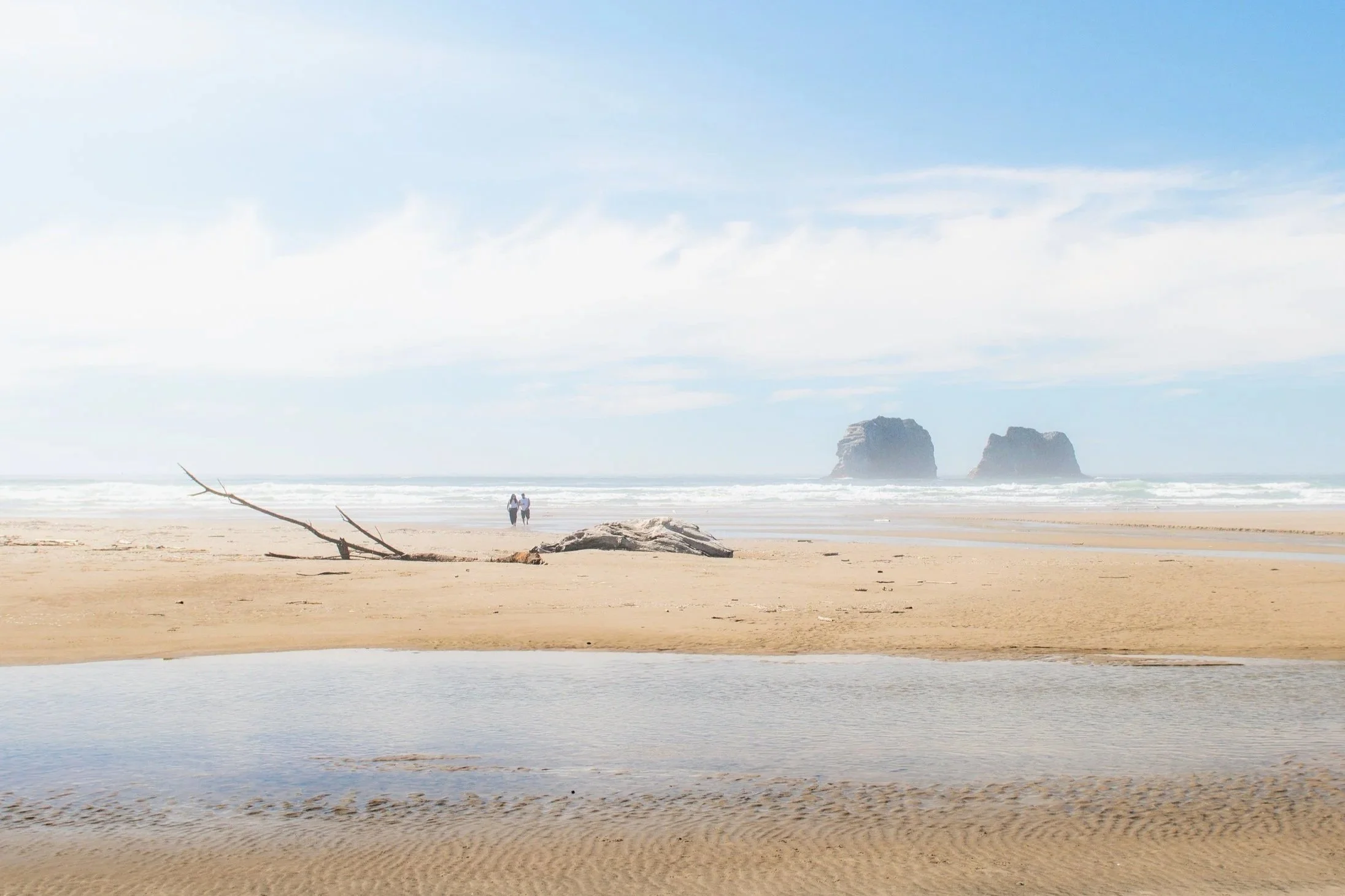 Sunny beach with two people walking near the water at Oregon's Rockaway Beach, large rocks off the coast, driftwood on the sand