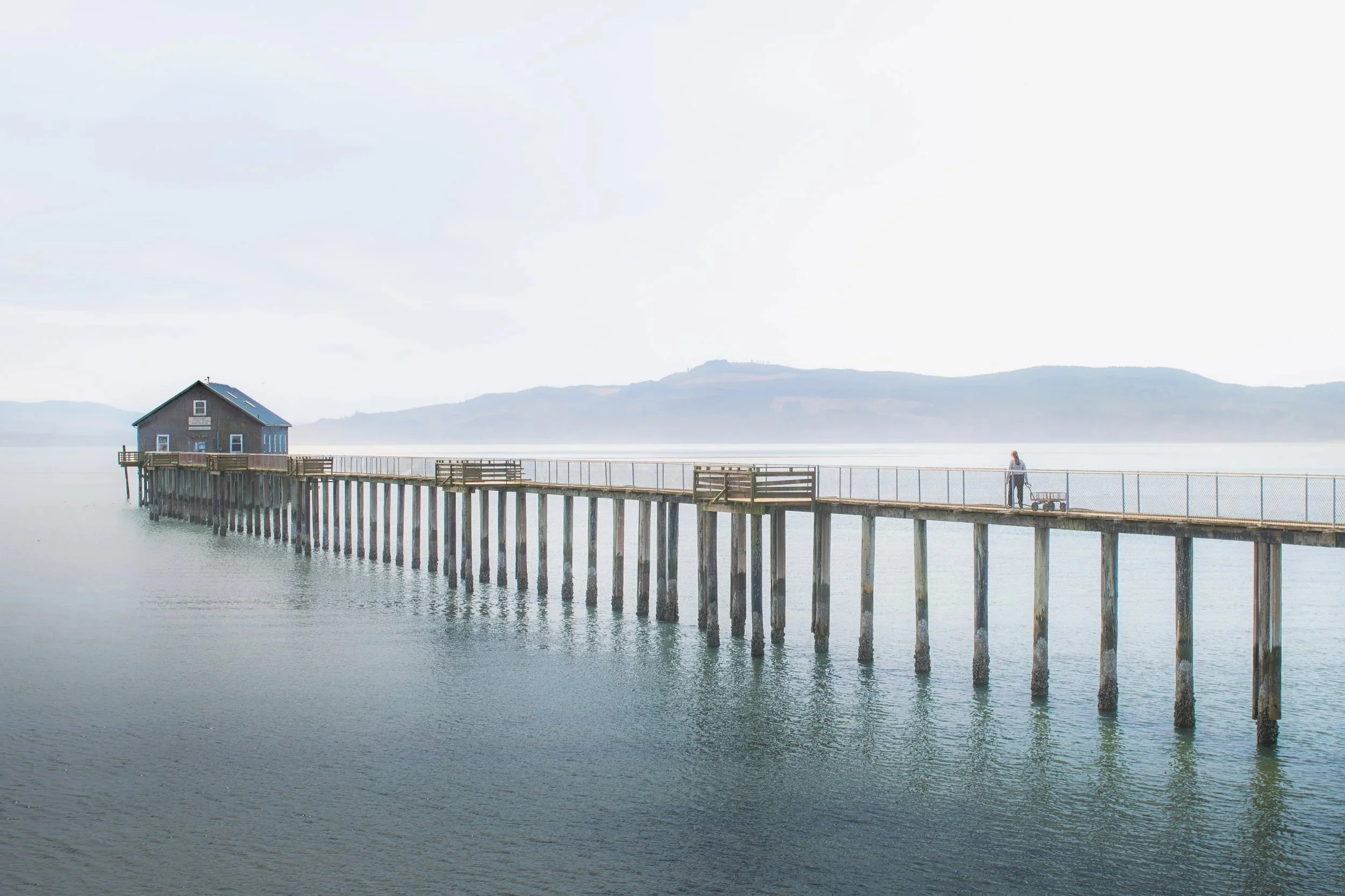 Travel experience photography- Oregon Coast A person walking on a pier over the water with a small house on stilts and a bench, against a backdrop of distant hills and a cloudy sky.
