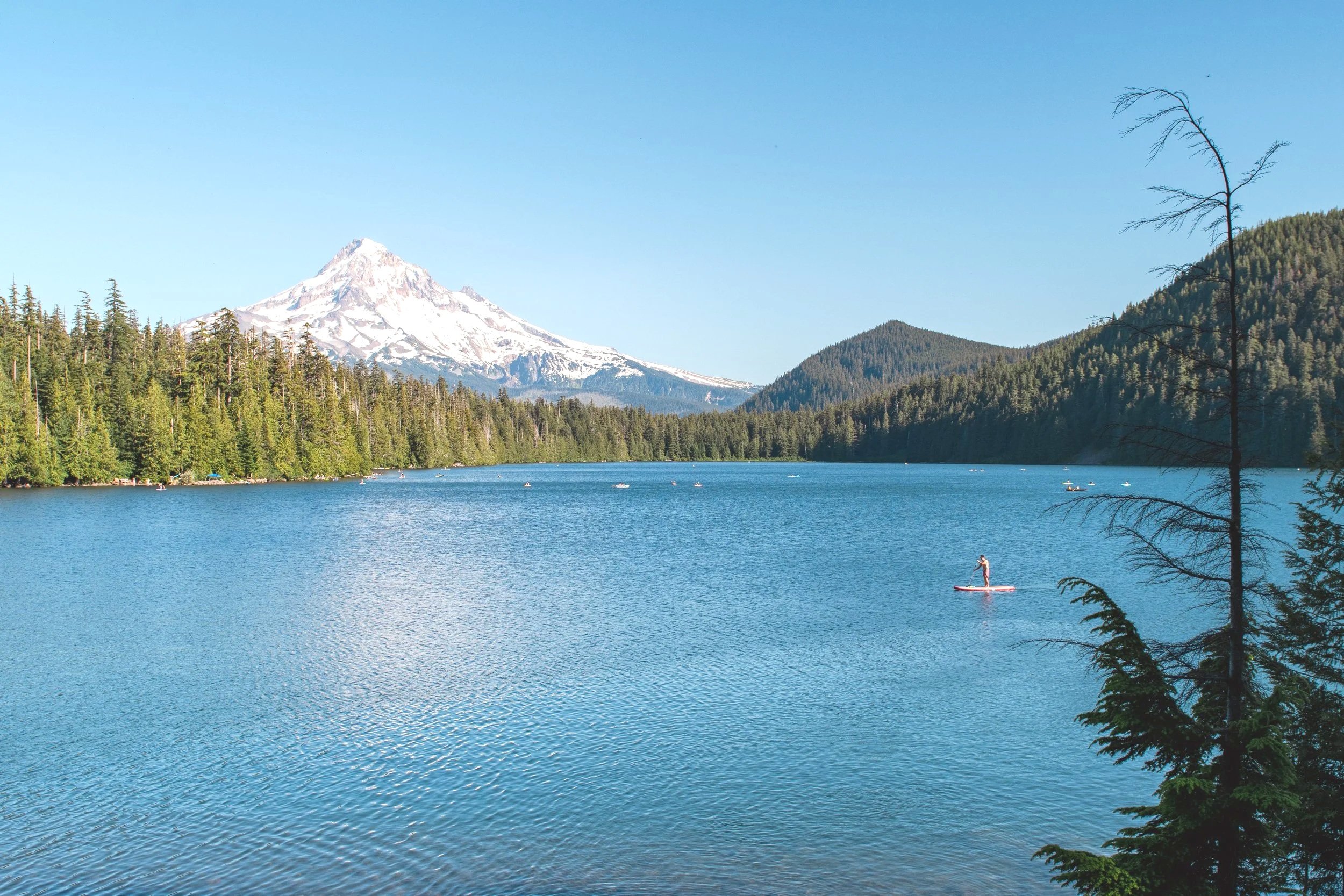  Oregon Travel- A scenic lake surrounded by forested mountains and a snow-capped Mt Hood in the background. A person paddleboarding on the lake.