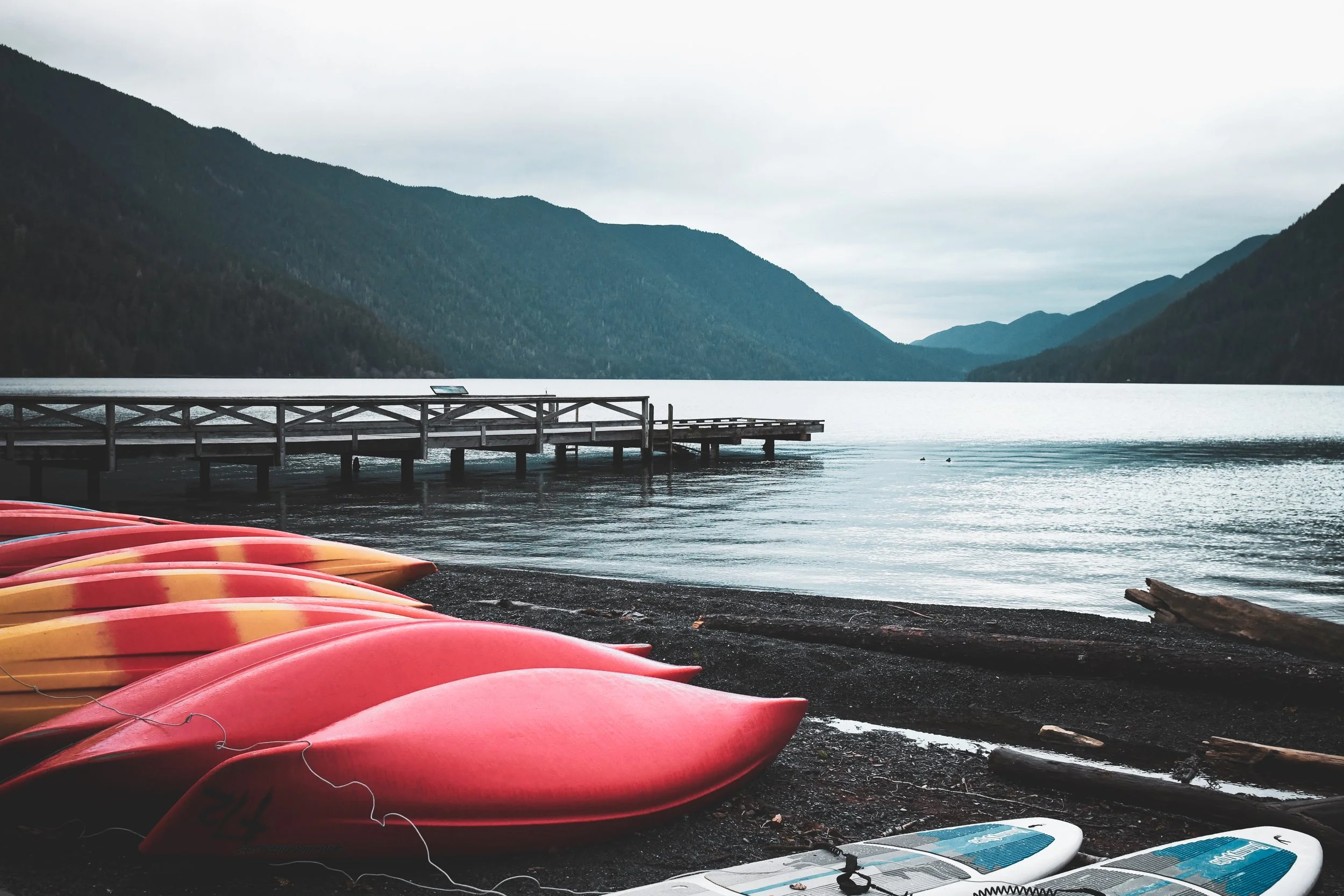 Row of red and yellow kayaks on a rocky shore with a wooden dock extending into Lake Crescent surrounded by mountains under an overcast sky.