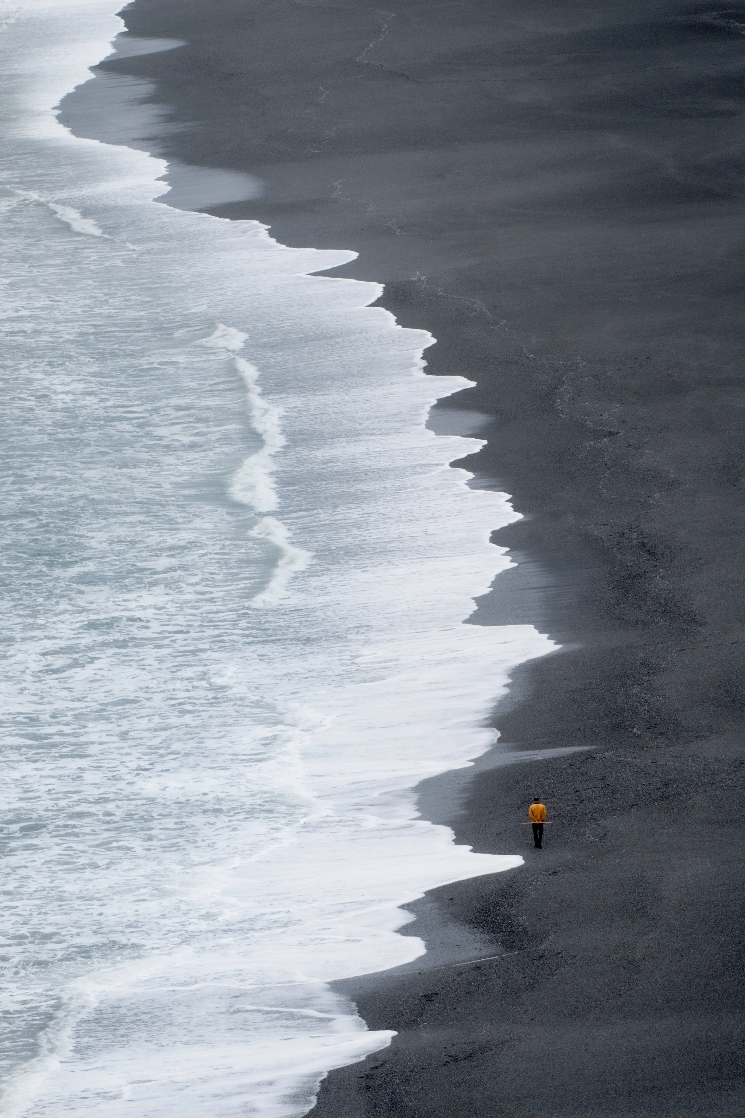 A person in a yellow jacket stands alone on a black sandy beach, observing the waves as they crash onto the shore, with the ocean extending into the distance.