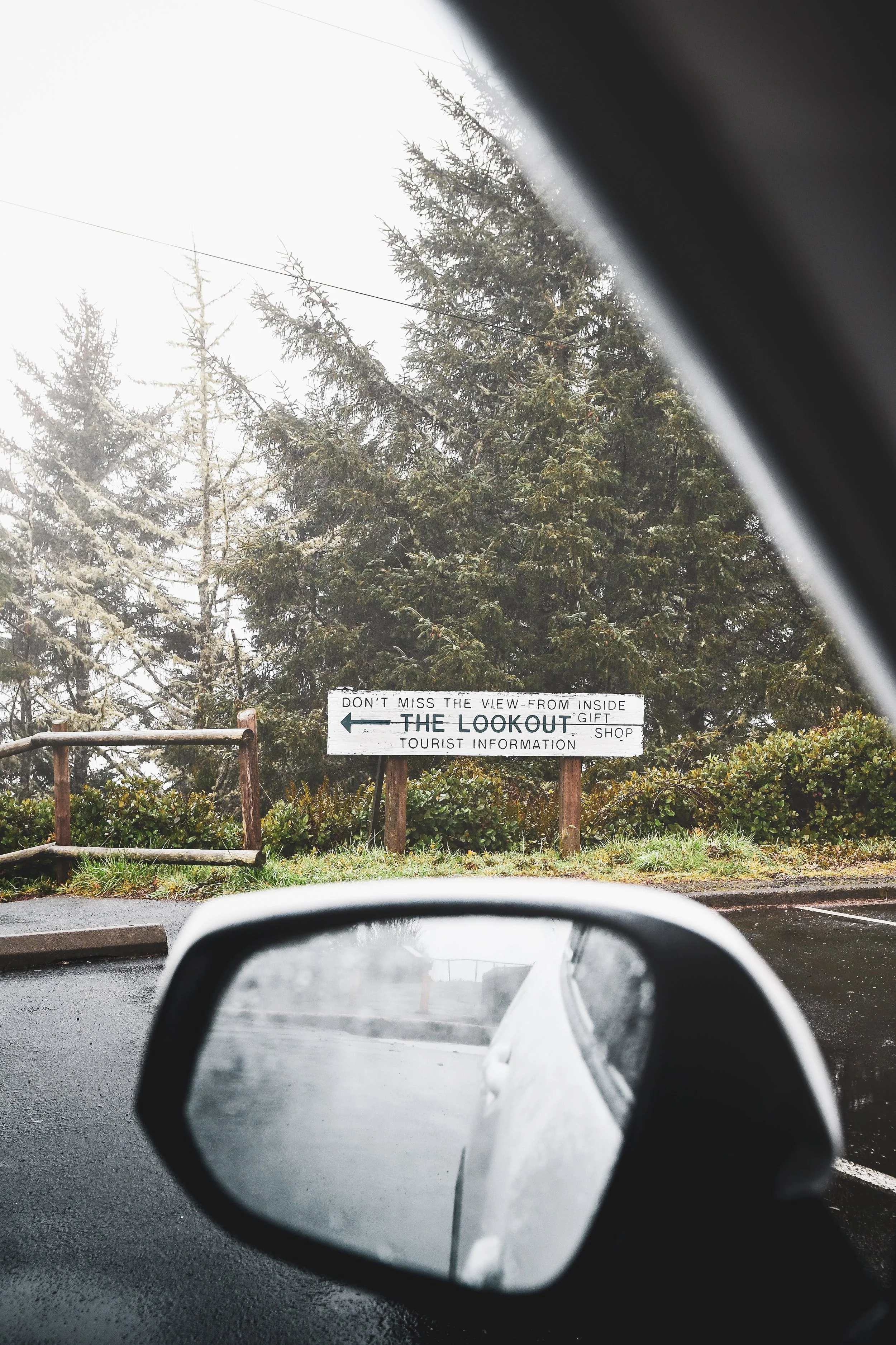 Travel experience photography- Oregon Coast View of a sign indicating a lookout point, with trees in the background and a car's side mirror in the foreground.