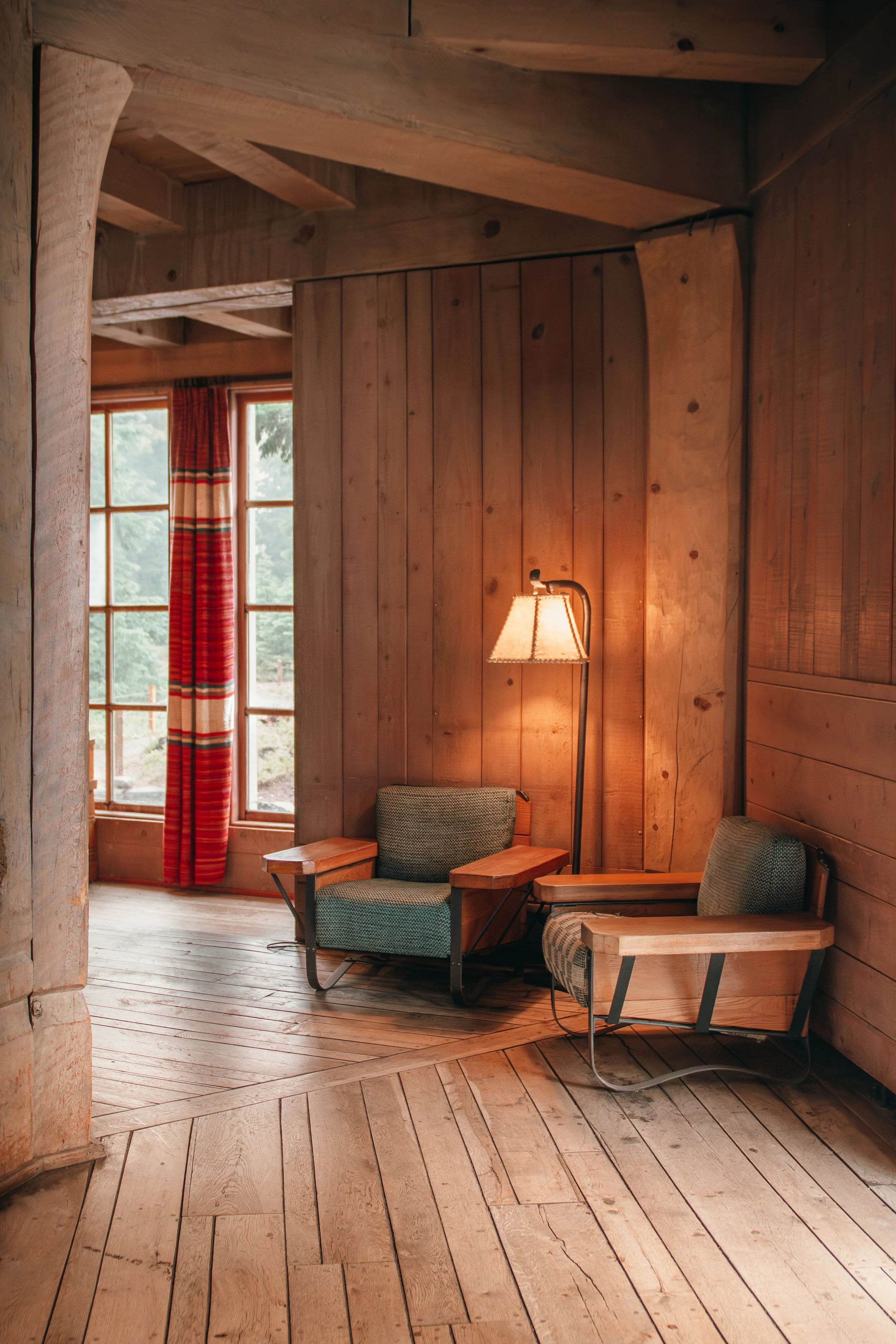 A cozy wooden room at Oregons timberline lodge with a large window, patterned curtains, and wooden furniture, including a chair with a small side table and a floor lamp.
