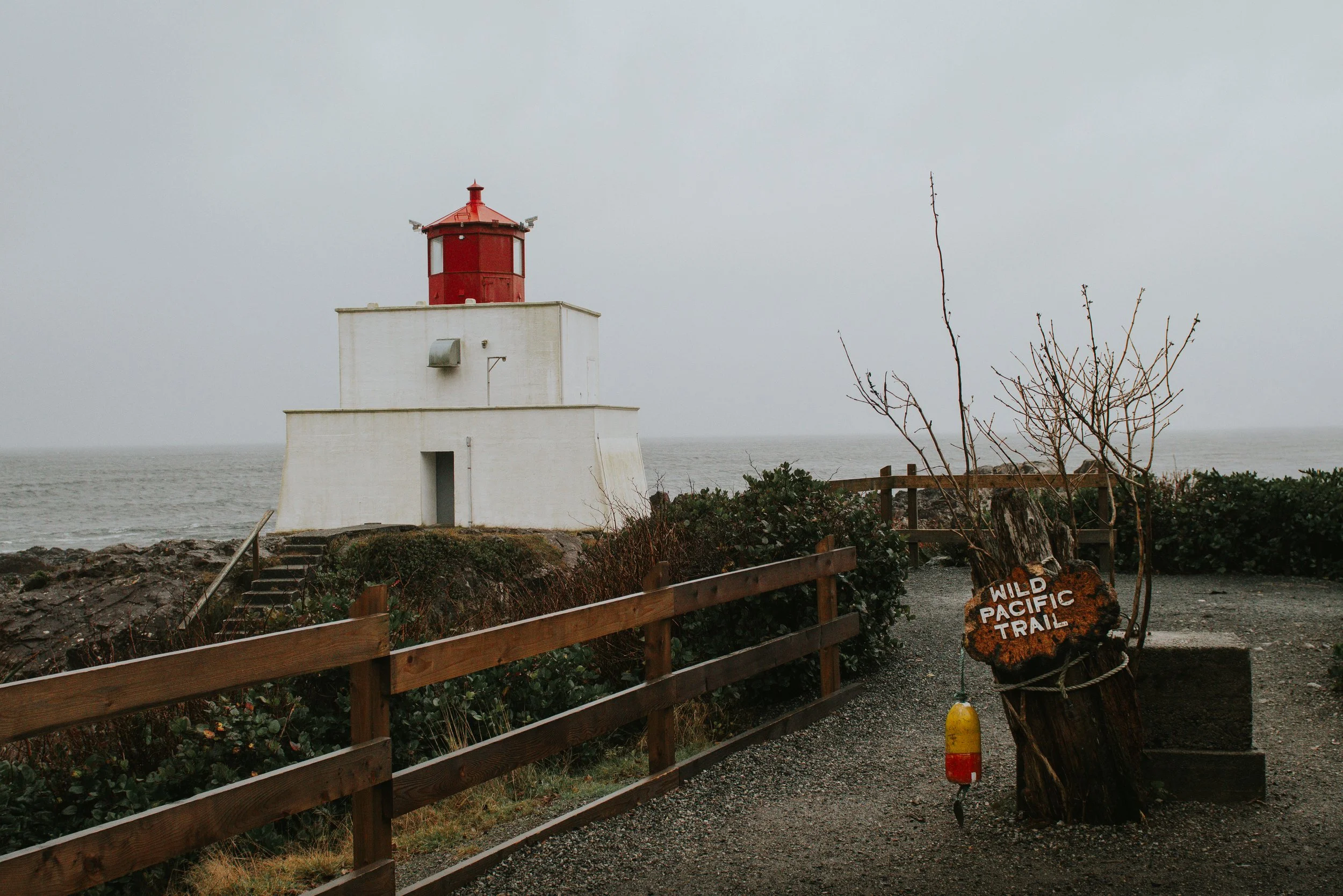 Travel Photography A white lighthouse on Vancouver Island with a red top located by the ocean, with a trail called 'Wild Pacific Trail,' a wooden fence, some trees, and a sign at the entrance, during cloudy weather.