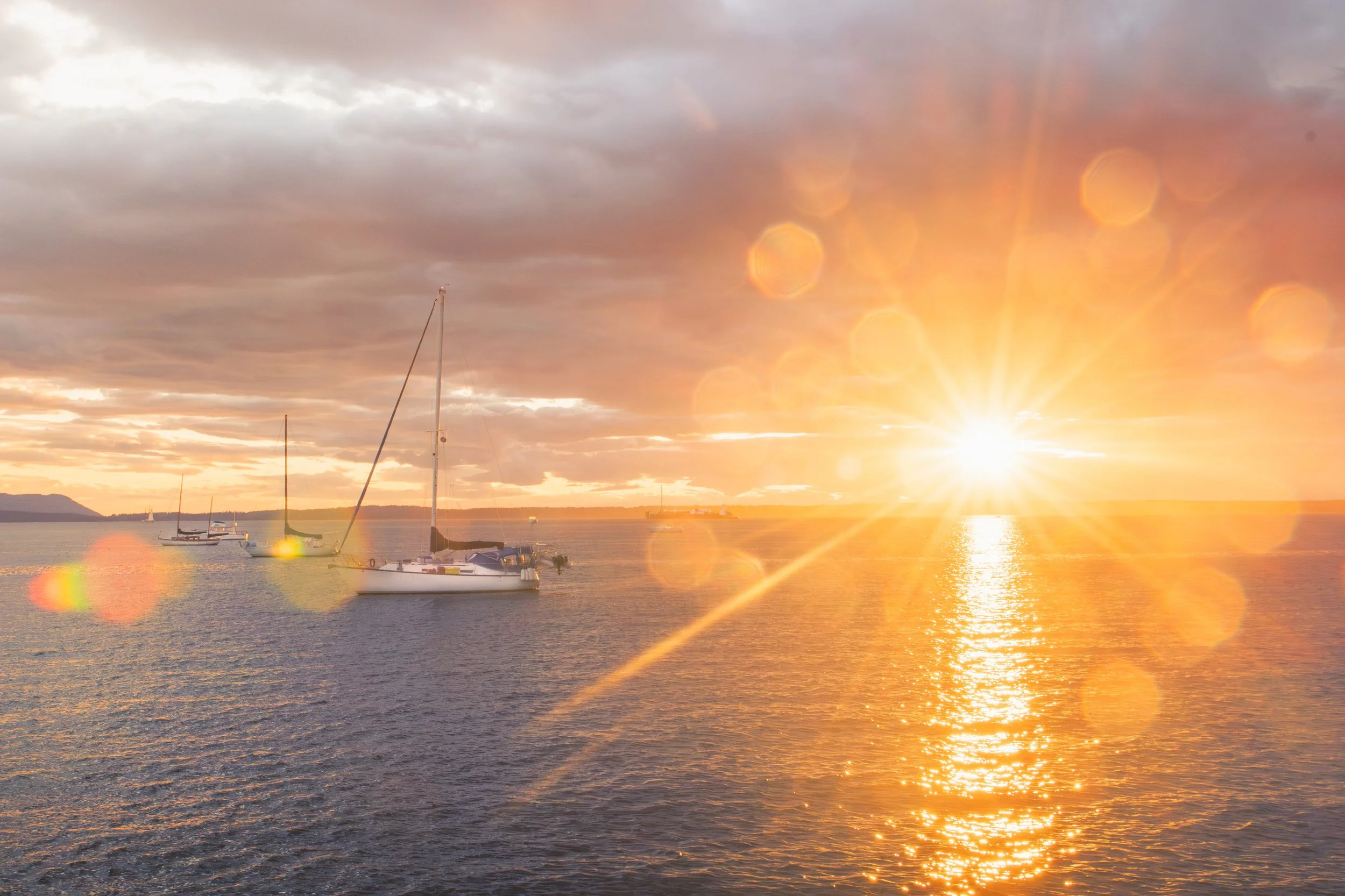 Bellingham travel activity-  sunset over a body of water with sailboats and a mountain in the distance, featuring lens flare and warm colors.