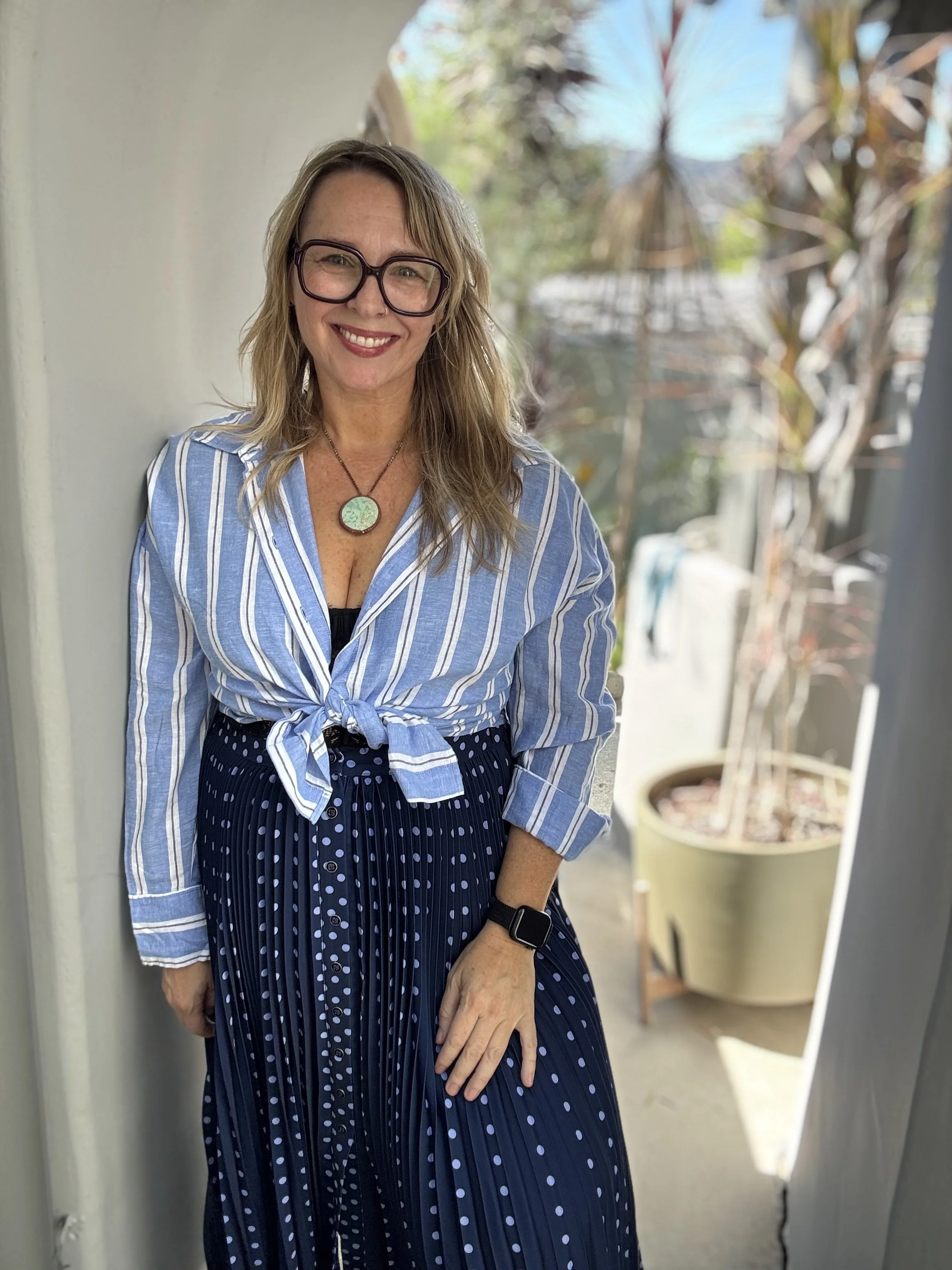 A smiling woman with blonde hair, glasses, and a necklace, standing outdoors near a white wall and potted plants, wearing a blue striped shirt and a navy polka-dot skirt.