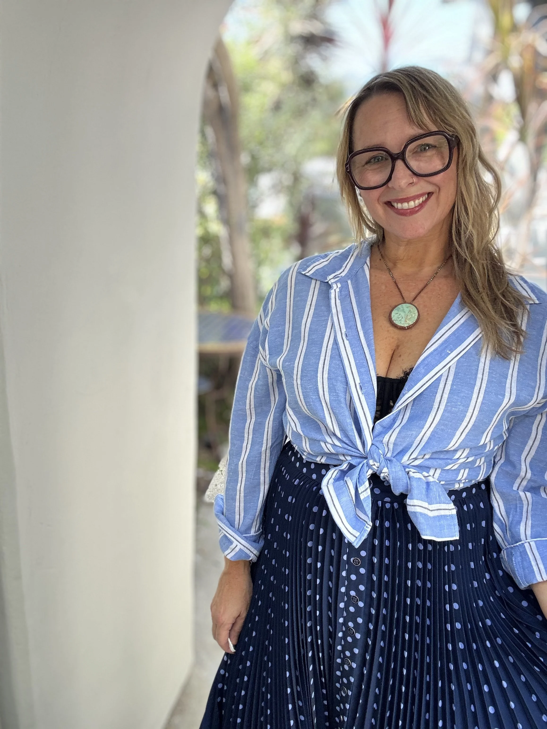 Smiling woman with glasses and wavy hair standing outdoors near a white wall, wearing a blue and white striped blouse and a navy skirt with white polka dots, with trees and sunlight in the background.