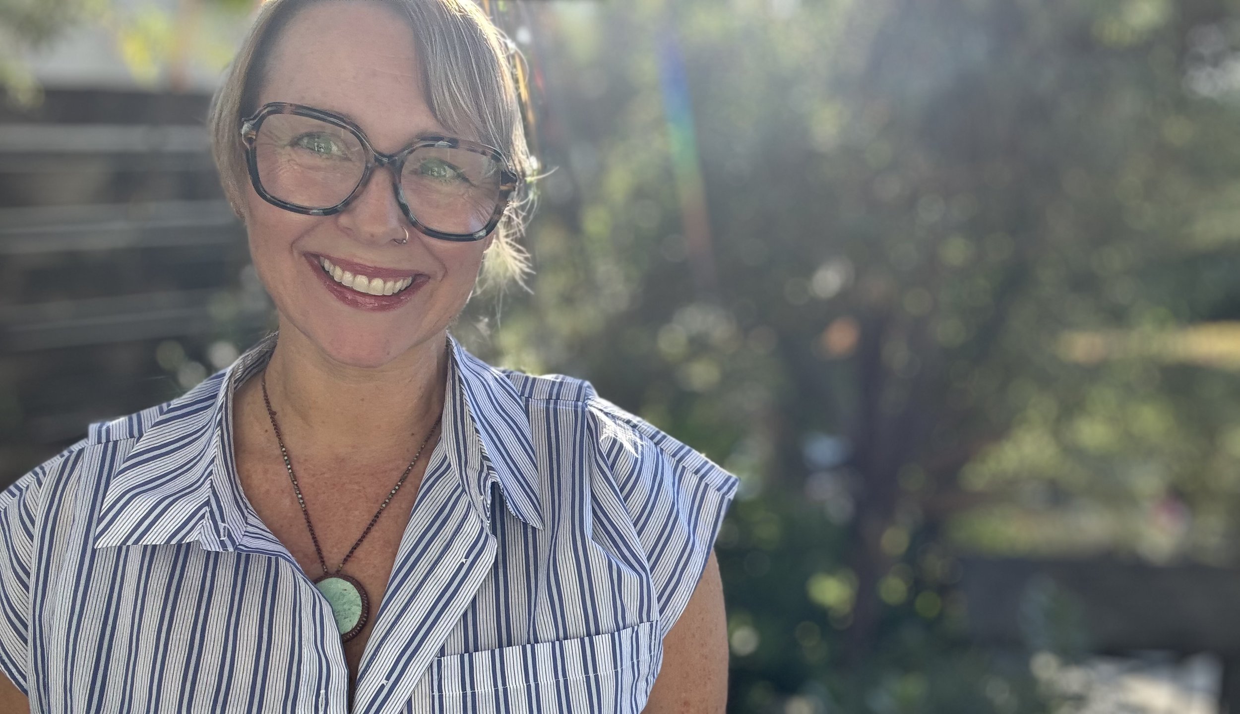 A woman with short hair wearing oversized glasses, smiling outdoors on a sunny day, with a background of trees and bokeh sunlight.
