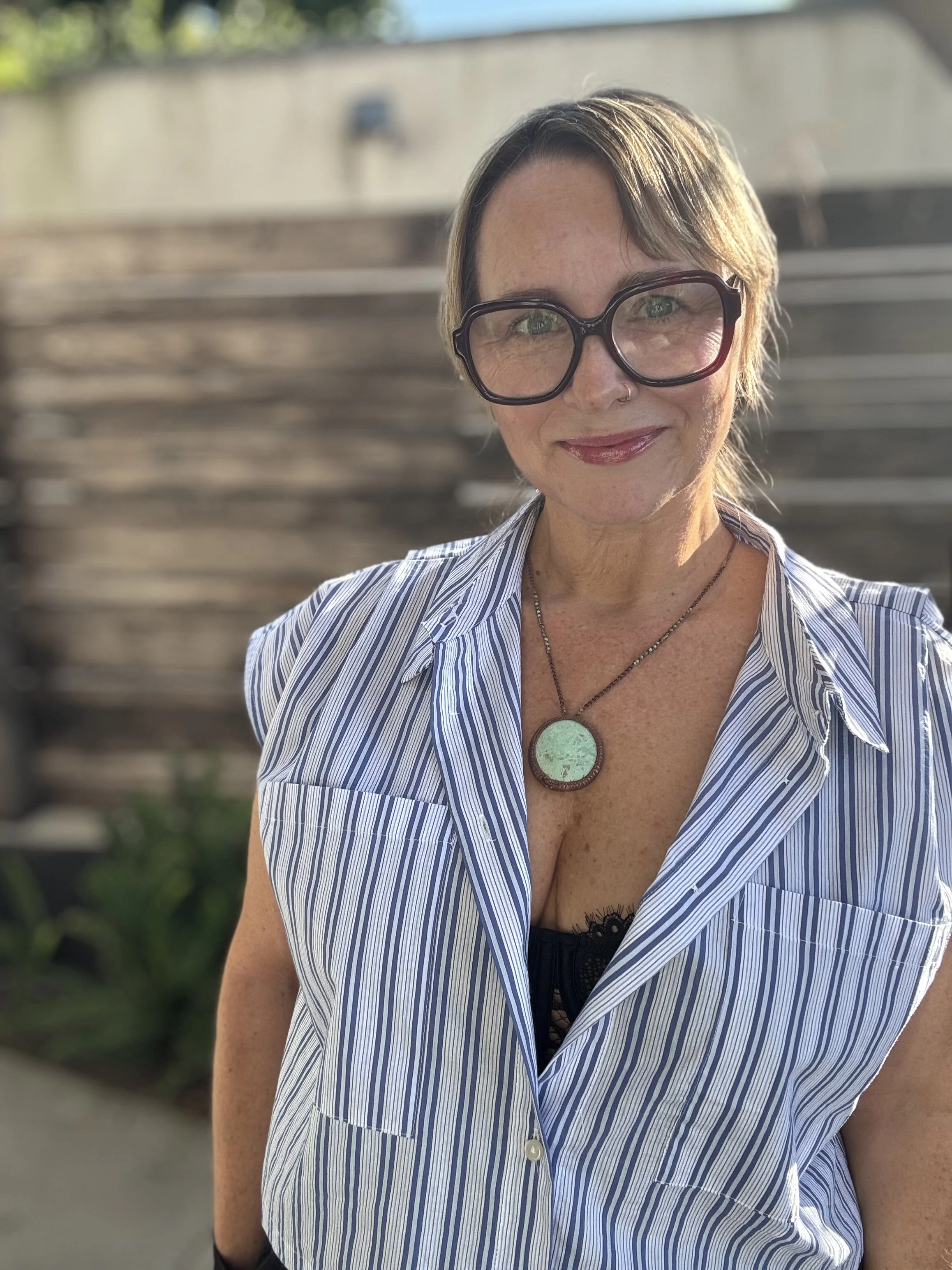 A woman with short brown hair wearing large black glasses, a striped blue and white shirt, and a green pendant necklace, smiling outdoors with a blurred background of stairs and greenery.