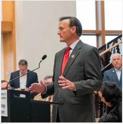 Man in a gray suit and red tie speaking at a podium in a formal setting, with several people seated behind him.