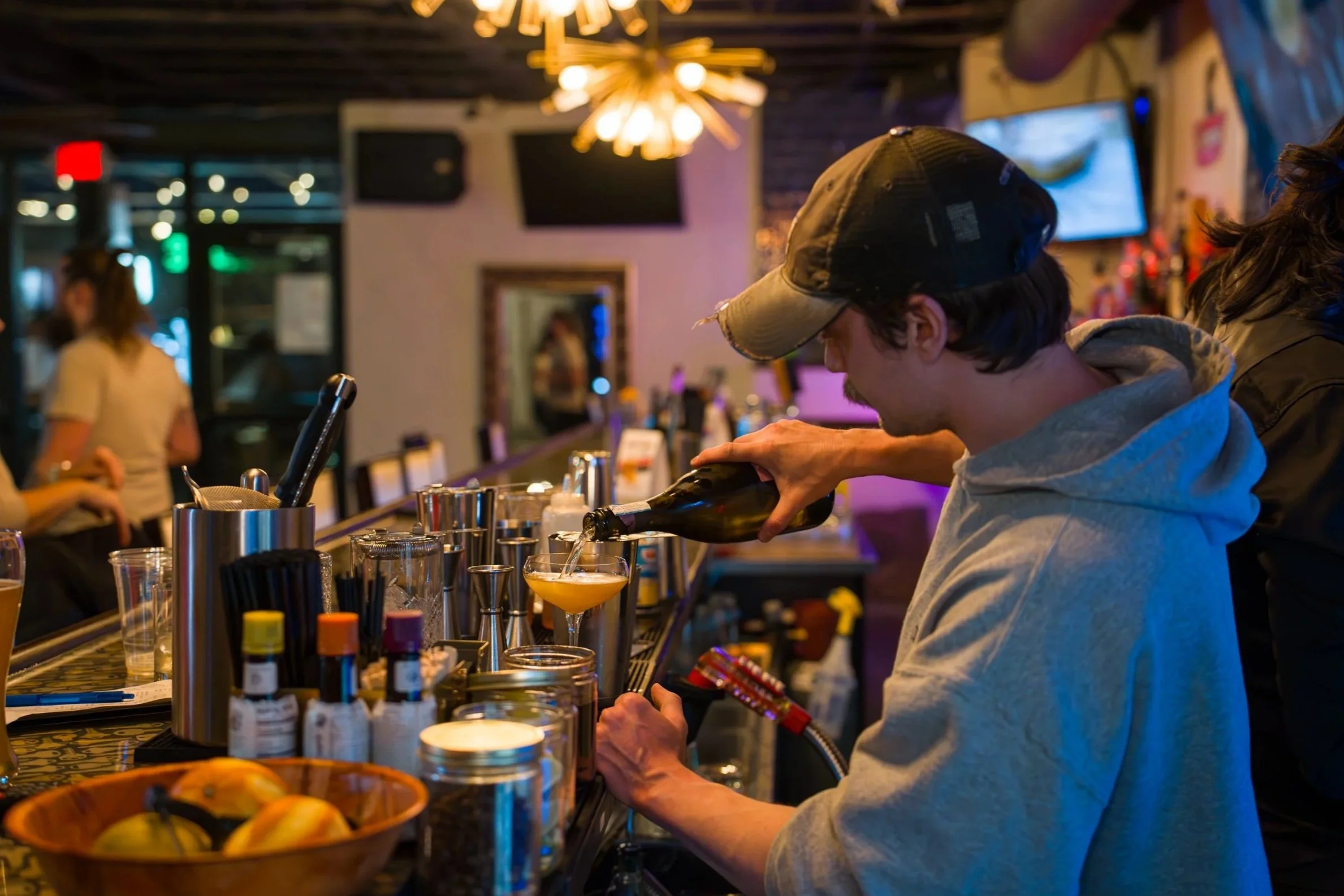 A bartender pouring a drink at a bar in a lively, dimly lit restaurant or bar.