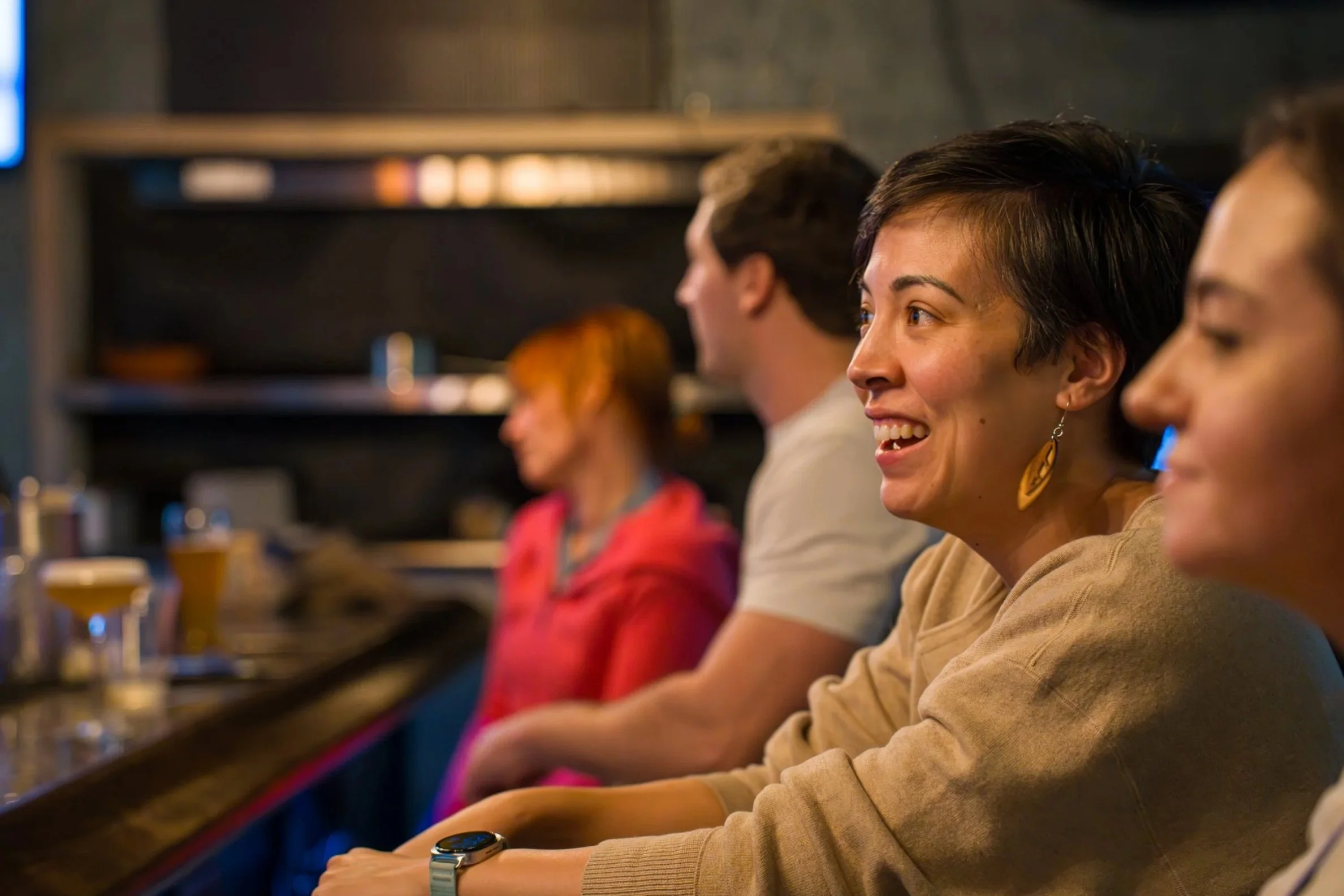 People sitting at a bar, smiling and engaging, with drinks on the counter, in a cozy, dimly lit setting.