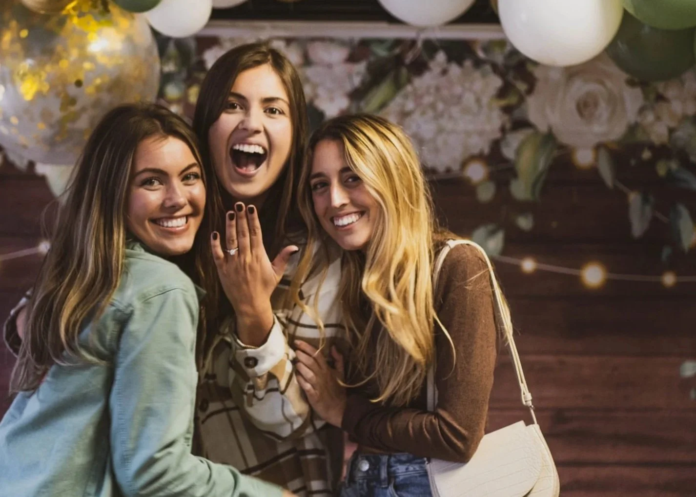 Three women celebrating and smiling at a gathering with balloons and floral decorations.