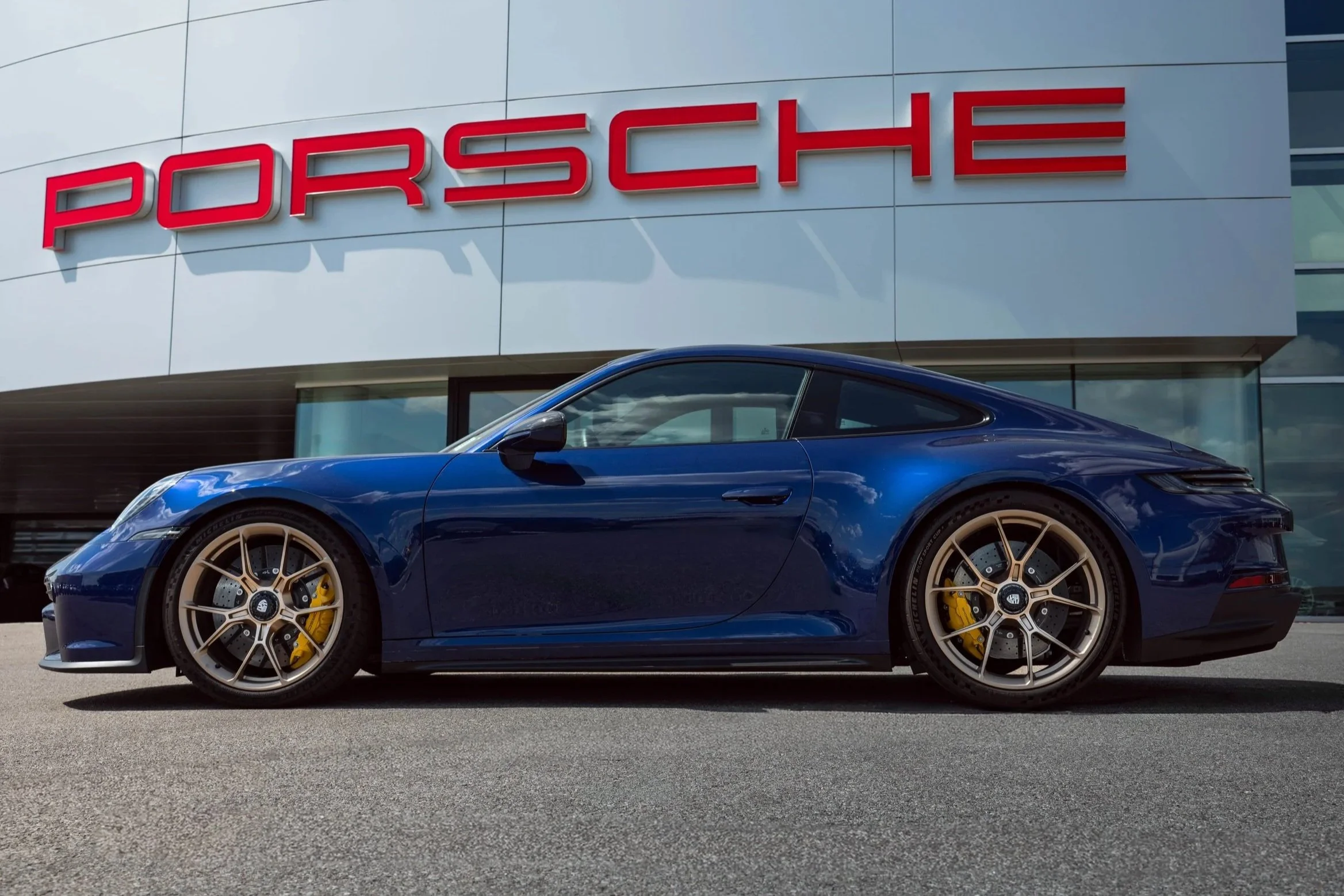 A blue Porsche sports car parked outside a dealership with a large red 'Porsche' sign on the building.