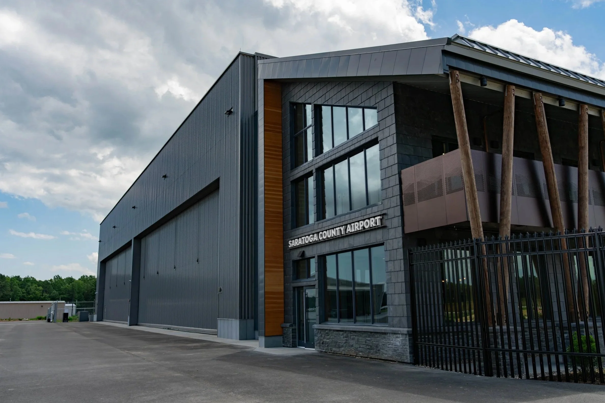 Saratoga County Airport with modern gray and stone building and large windows, surrounded by a black fence and parking lot under a partly cloudy sky.