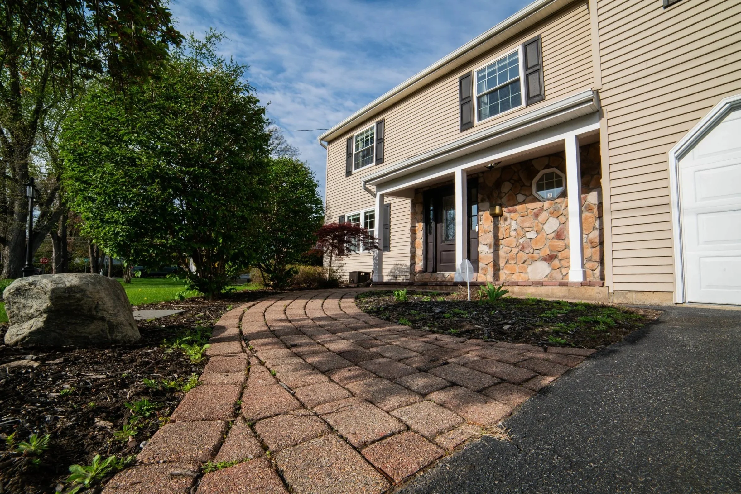 Exterior view of a house with a curved brick walkway leading to the front door, surrounded by greenery, trees, and lawn, under a partly cloudy sky.